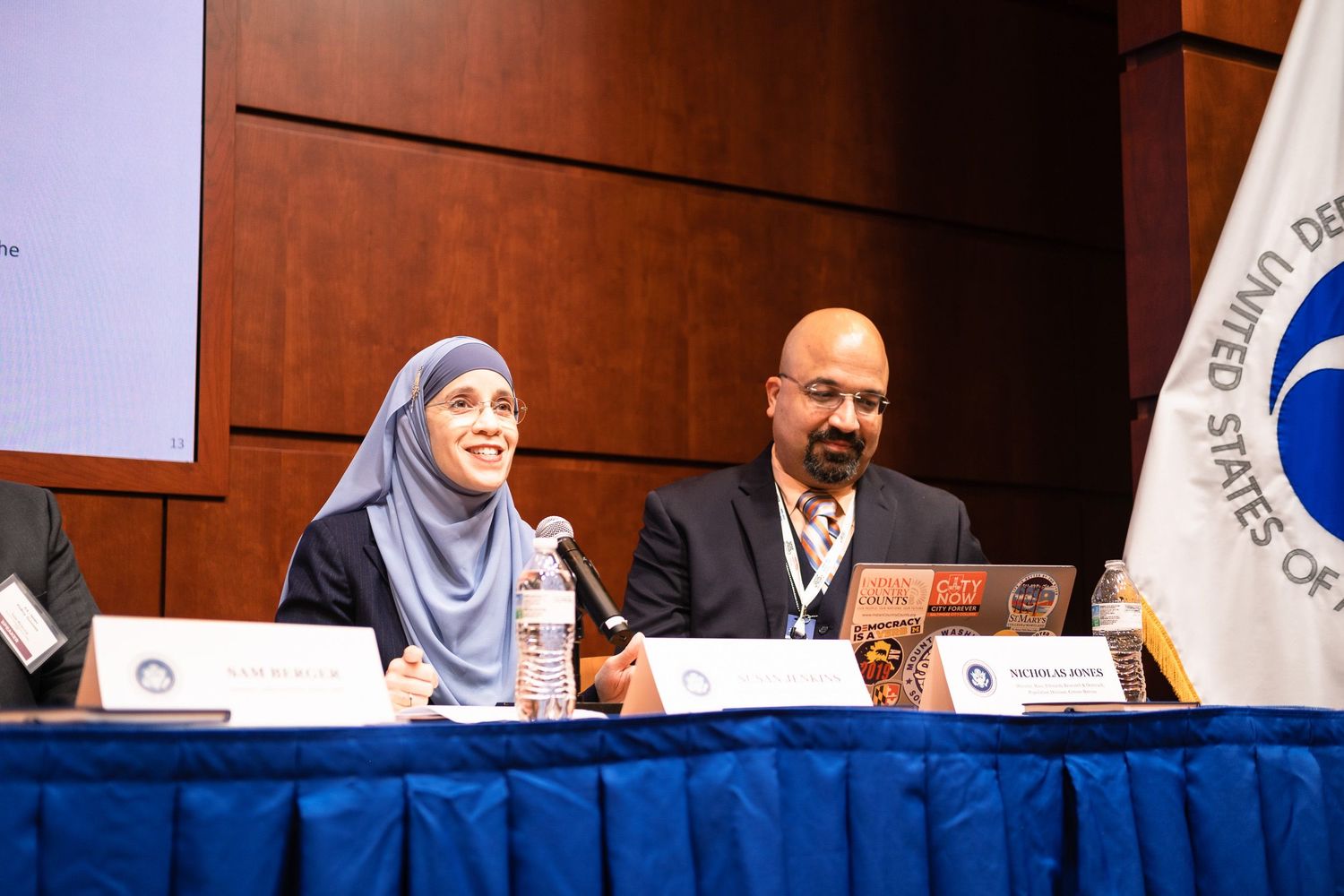 Two panelists, a woman in hijab and a man, seated at a conference table at a United States government event.
