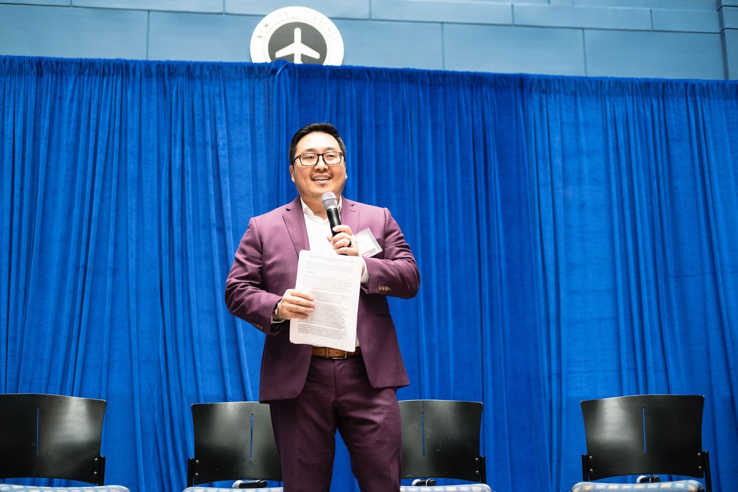 A smiling man in a purple suit holds papers and speaks into a microphone at a formal event with blue curtains.