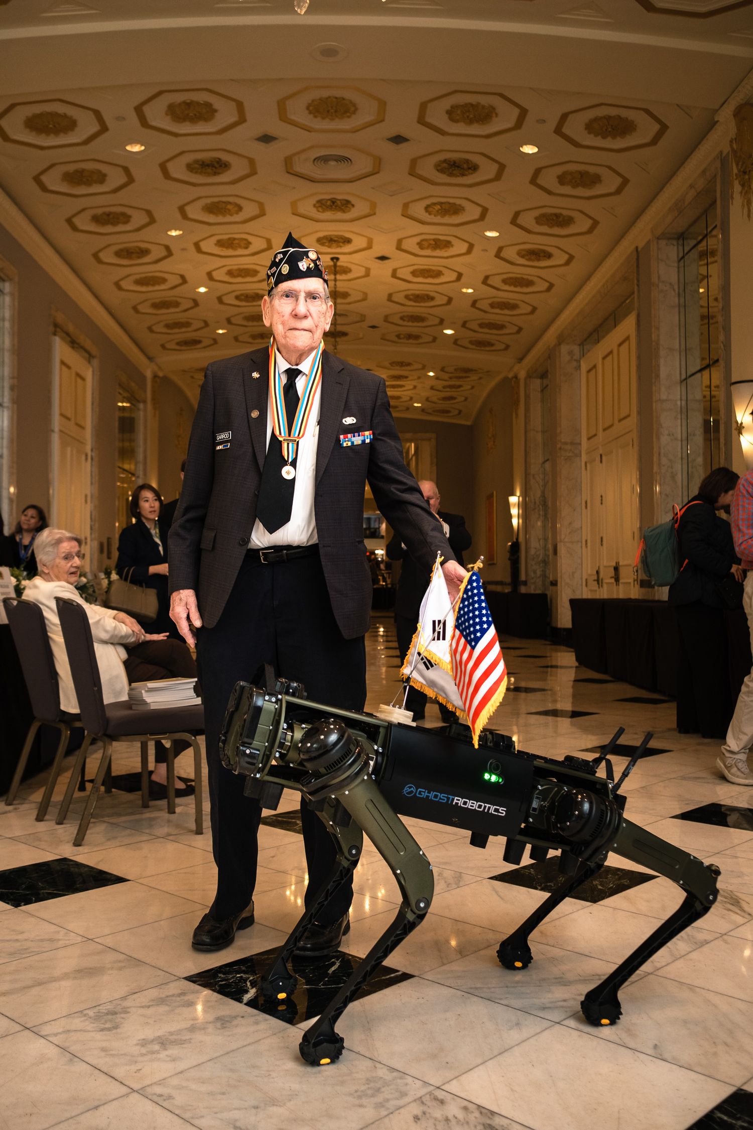 Veteran in military uniform stands proudly beside a Ghost Robotics quadruped robot dog carrying US and Korean flags indoors.