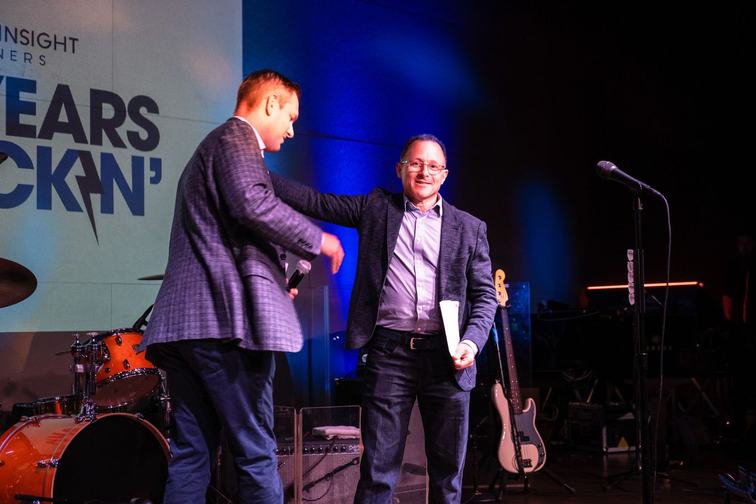 Two men shaking hands on stage at a corporate anniversary event with a rock-themed backdrop.