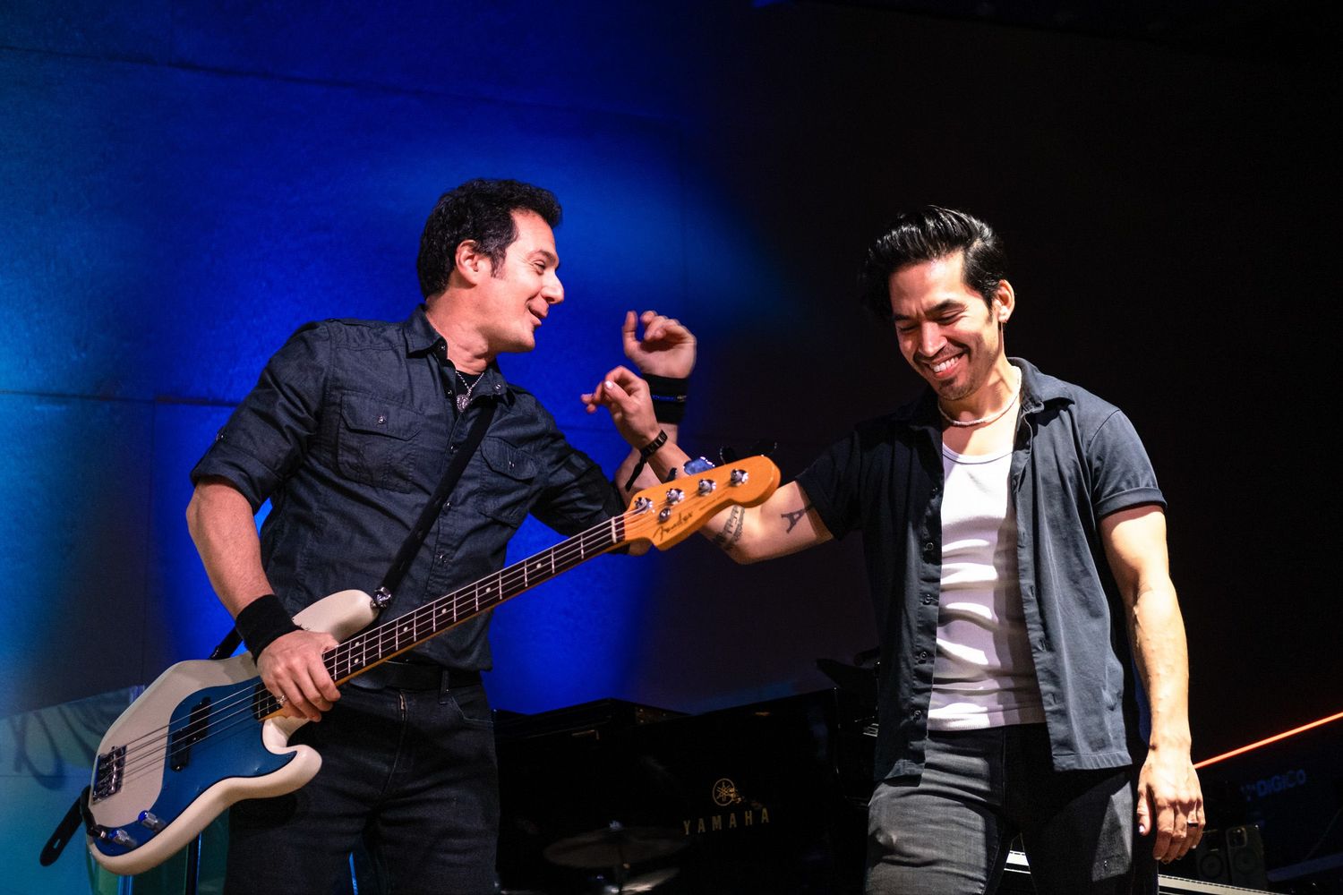Two male musicians share a joyful moment on stage, one holding a bass guitar under blue stage lighting.