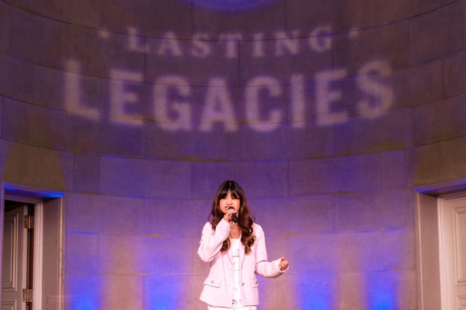 Young woman in white suit performing on stage at Lasting Legacies event with dramatic blue lighting backdrop.