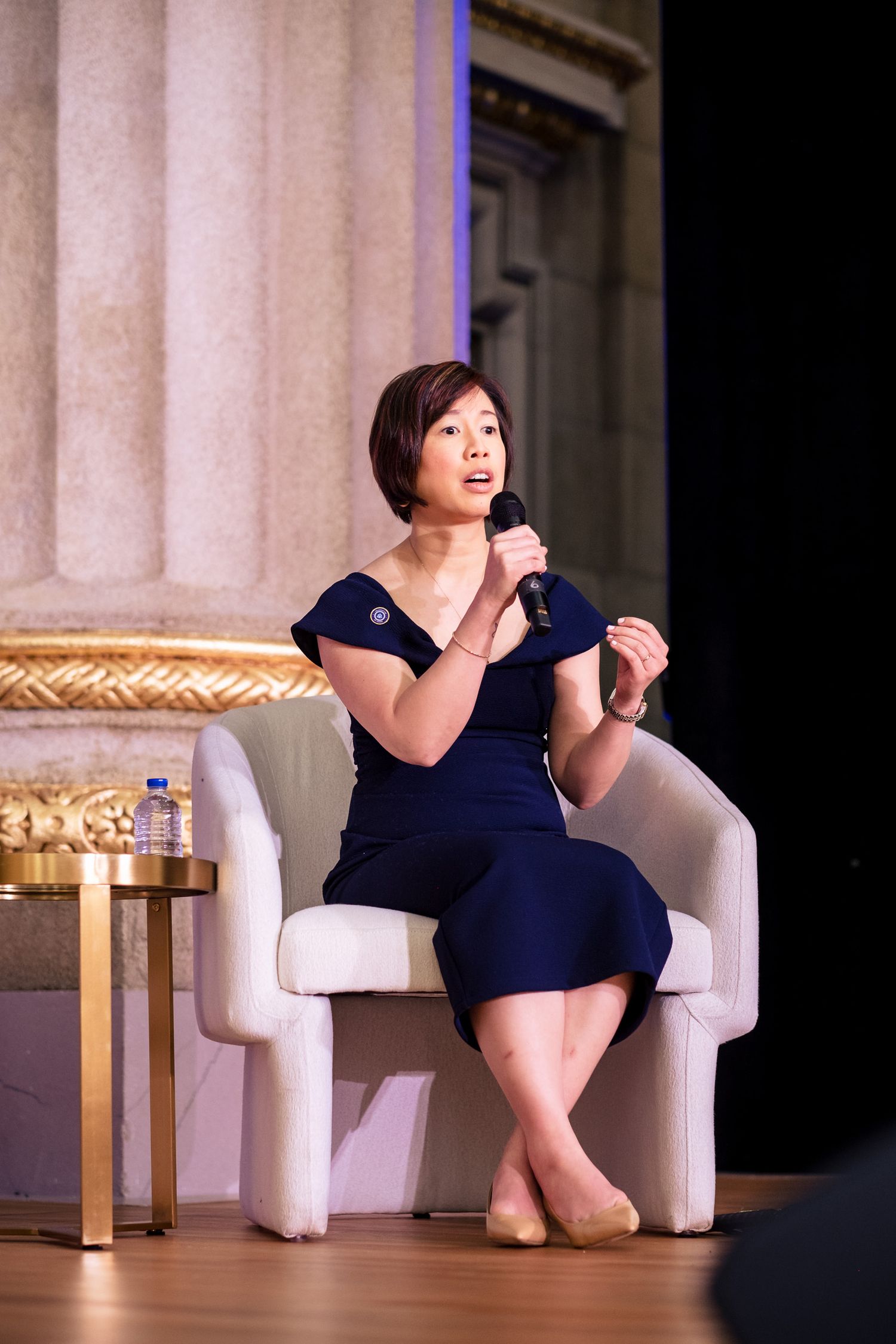 Woman in navy dress speaks into microphone while seated on white chair at elegant event.