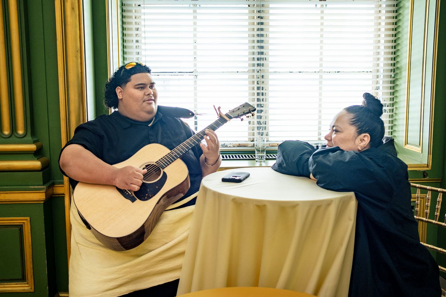 A man playing acoustic guitar serenades a woman at a table in an elegant green-walled room.