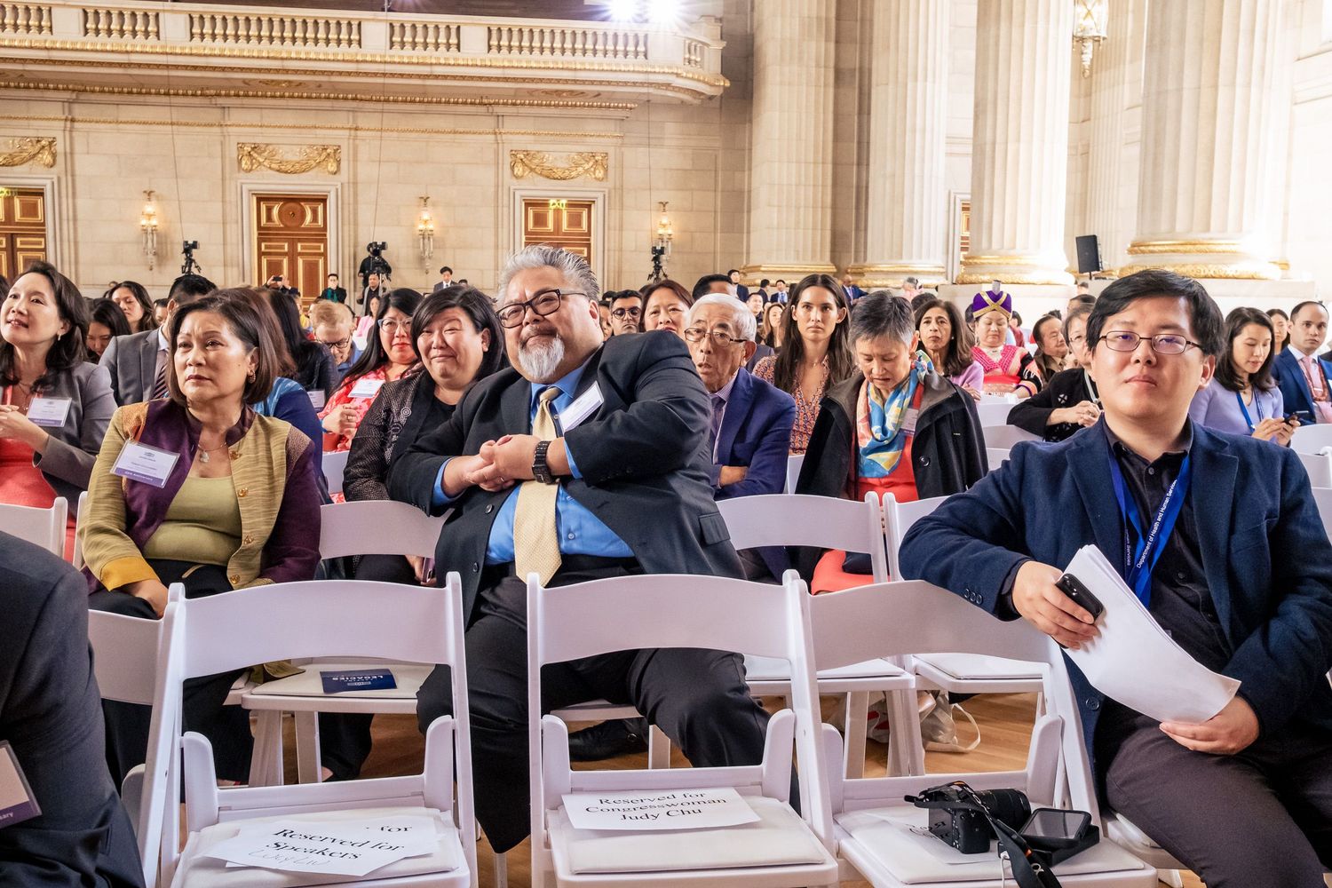 Diverse audience seated in elegant hall at a conference, smiling and engaged during a presentation.