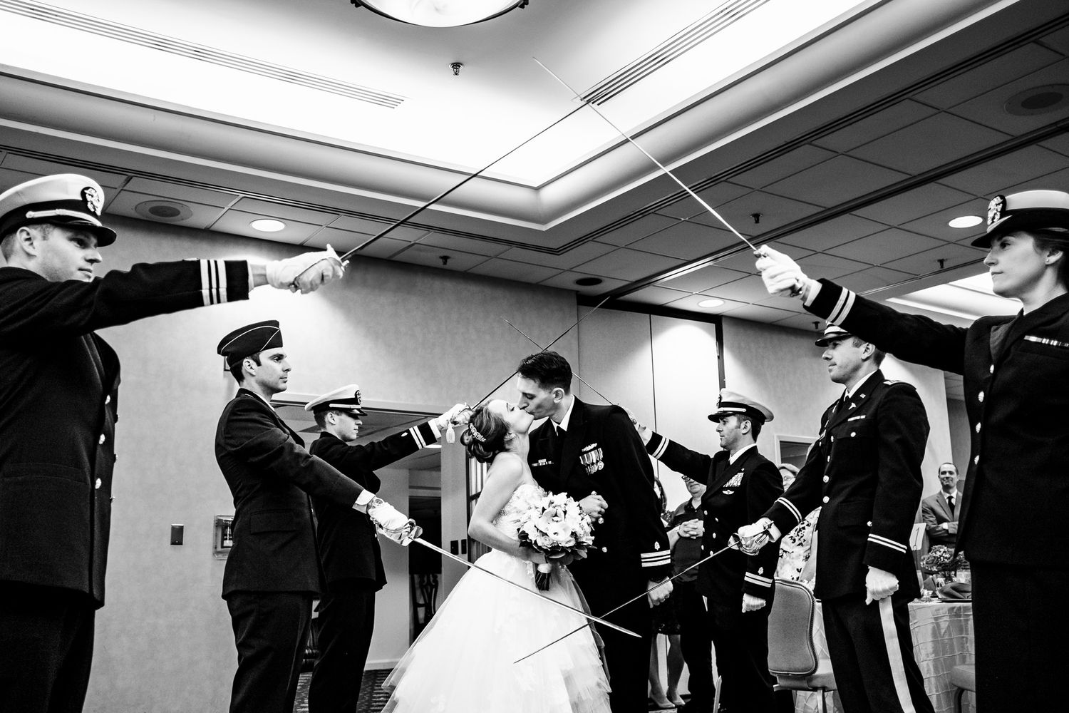 Military wedding couple kisses under an arch of swords formed by uniformed officers indoors.