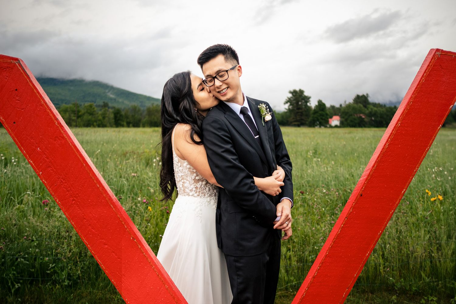 Bride and groom embrace romantically between large red letter V sculptures in a scenic mountain meadow.