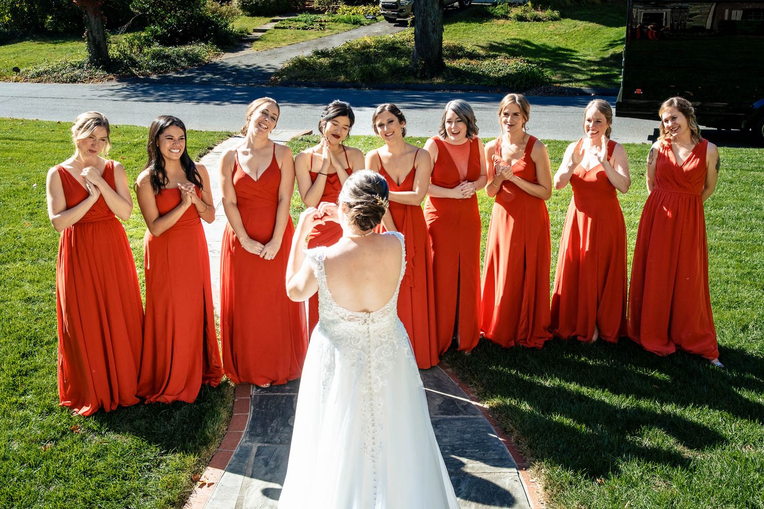 Bride reveals her gown to ten bridesmaids in red dresses on a green lawn.