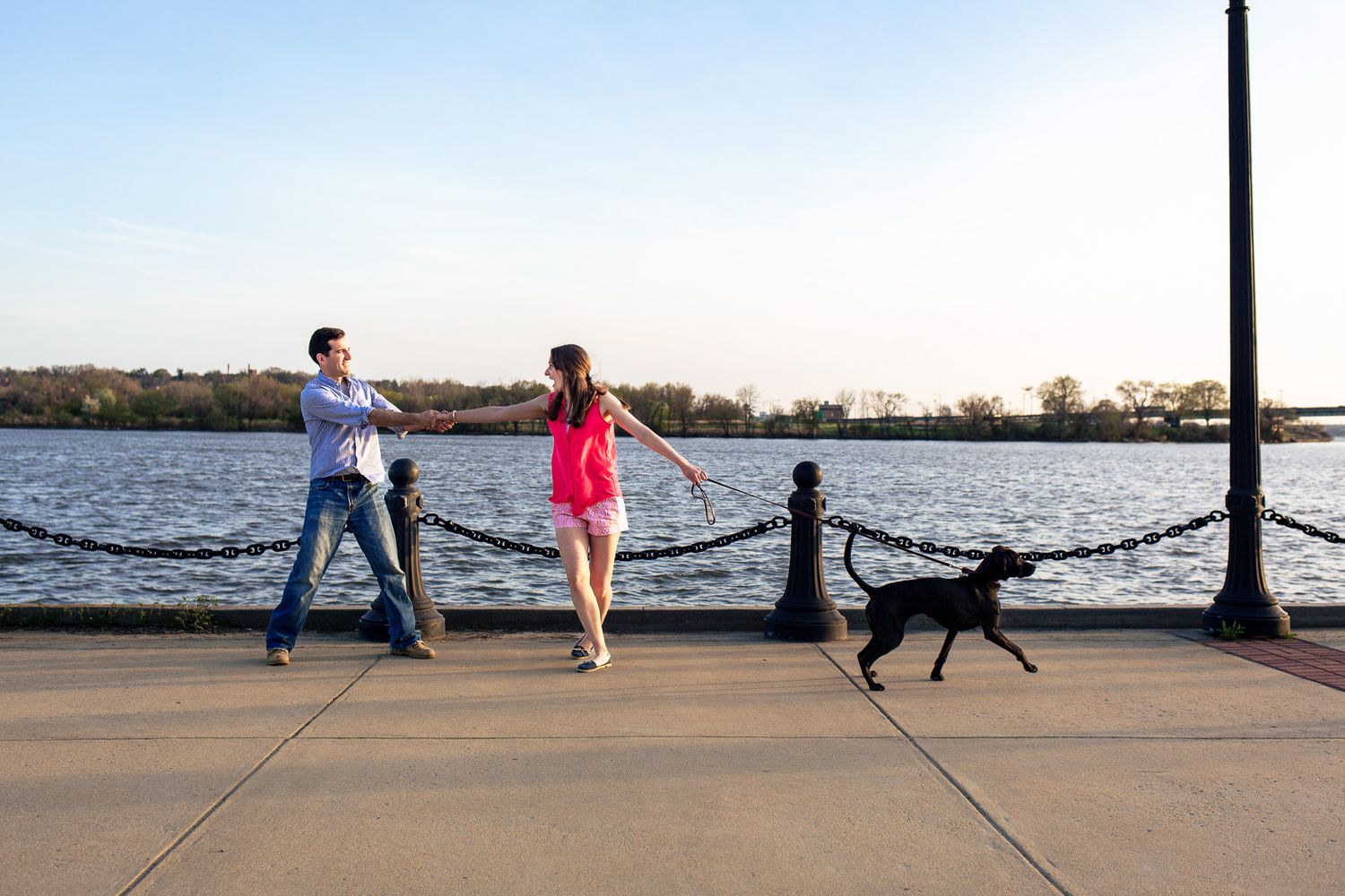 Couple walking a black dog along a scenic waterfront promenade on a clear sunny day.