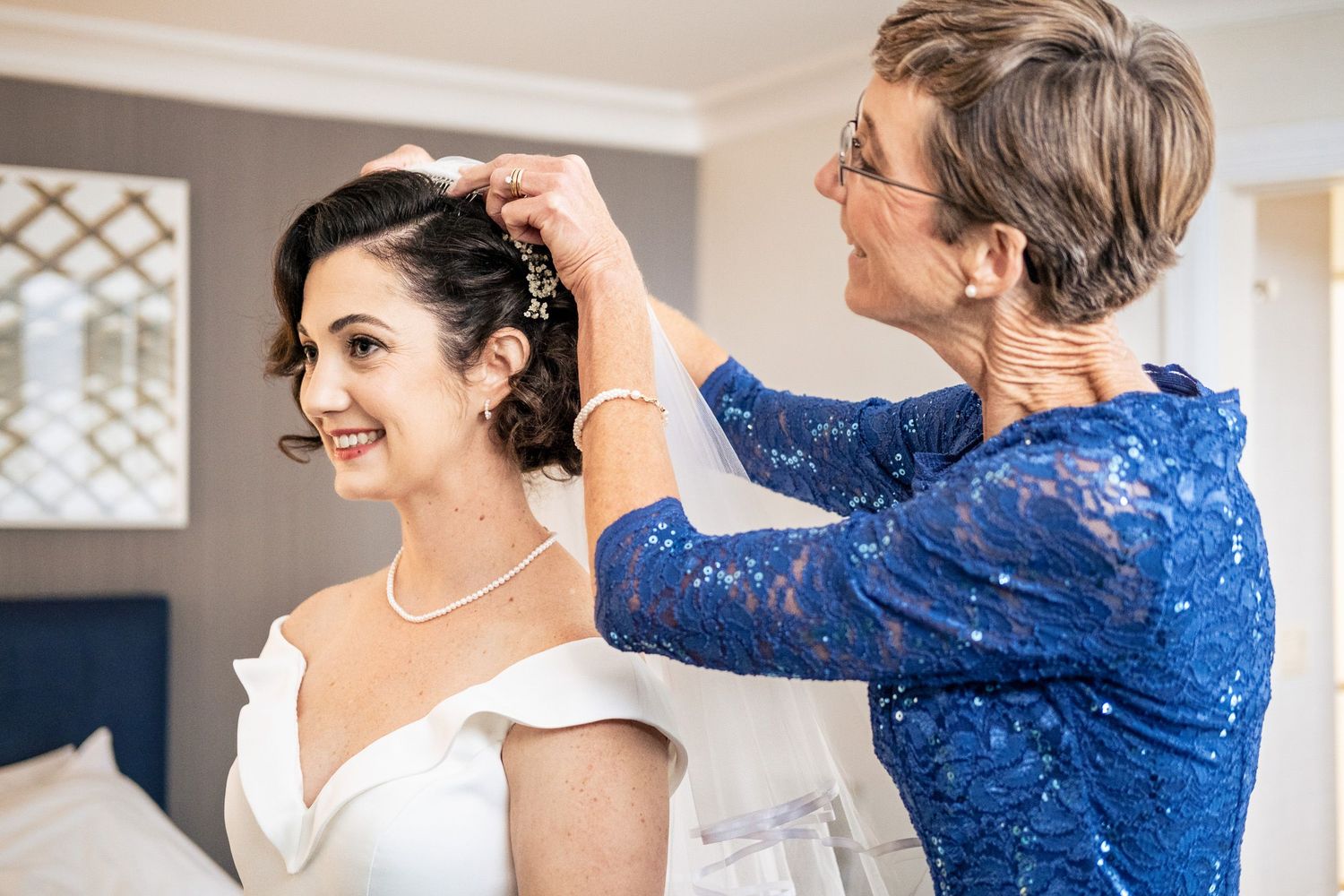 Smiling bride in white gown has hair accessory placed by older woman in blue lace dress.