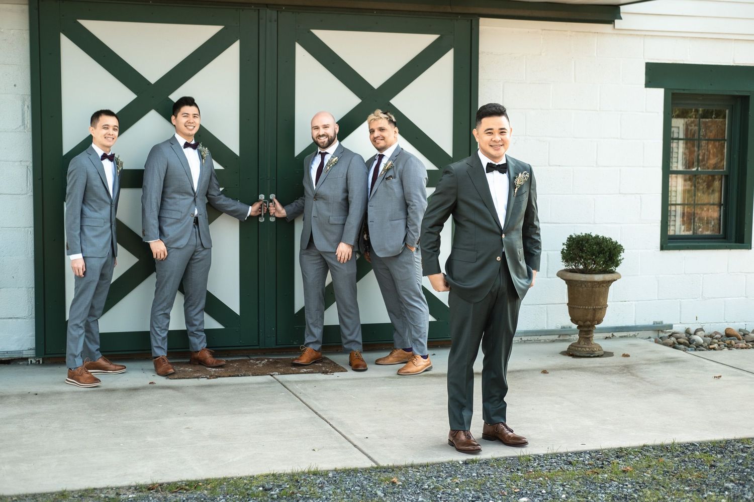 Four groomsmen in gray suits with black bow ties posing outside green barn doors.