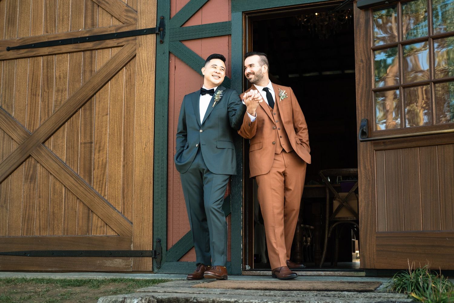 Two grooms stand hand in hand in the doorway of a rustic barn wedding venue.