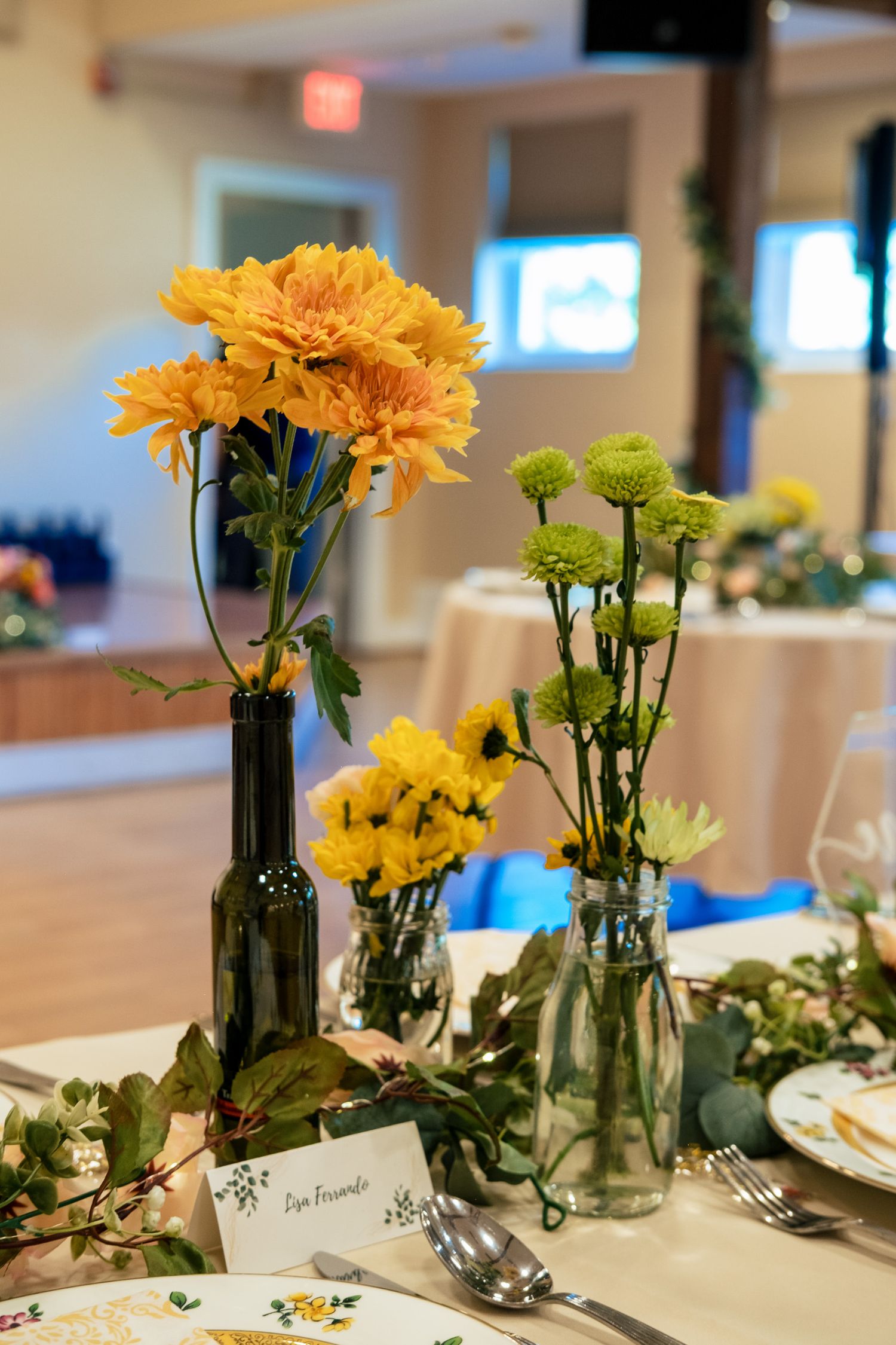 Elegant wedding table centerpiece with yellow chrysanthemums and green flowers in glass bottles and vases.