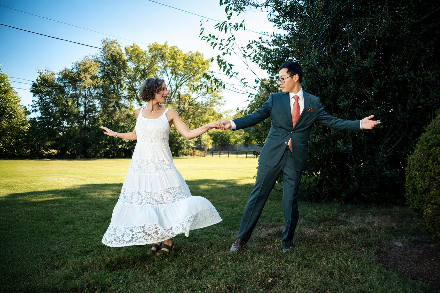 Joyful couple playfully spinning and holding hands on a sunny green lawn during their wedding day.