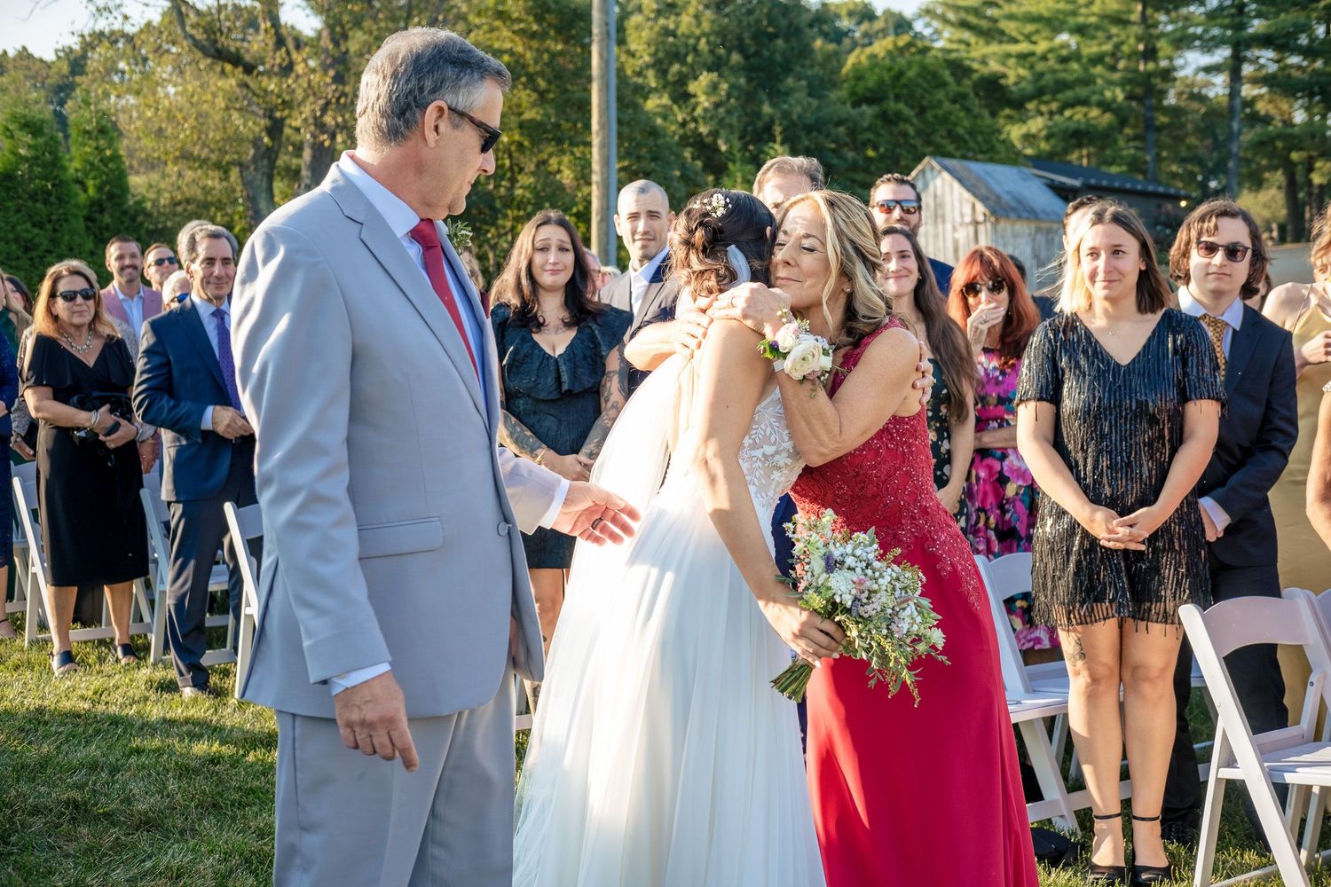 Bride shares an emotional hug with a woman in a red dress while a man in a gray suit looks on at the ceremony.