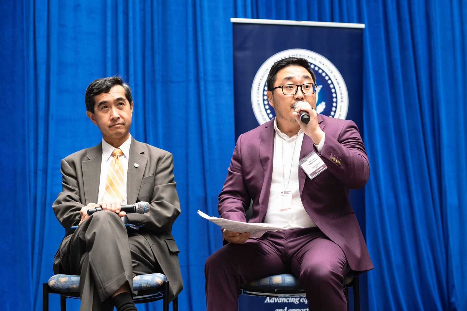 Two men in suits sit on stage during a formal panel discussion at an official government event.