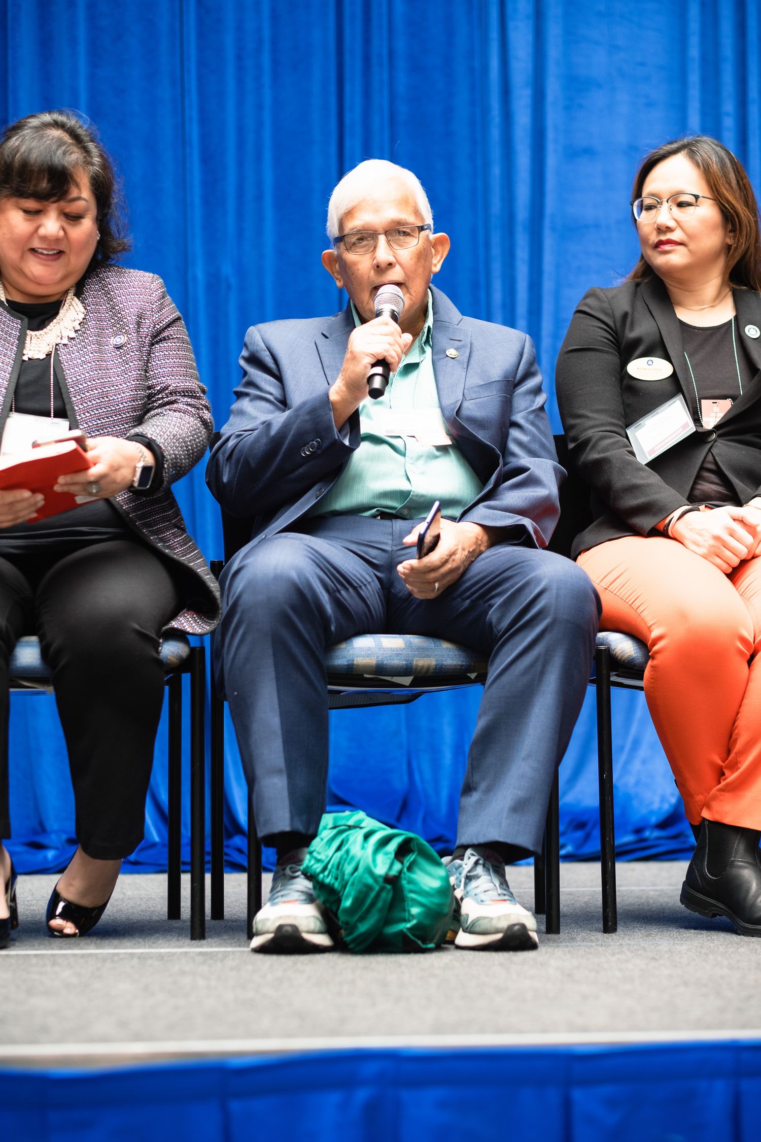 Three panelists seated on stage at a conference, with a man in a blue suit speaking into a microphone.