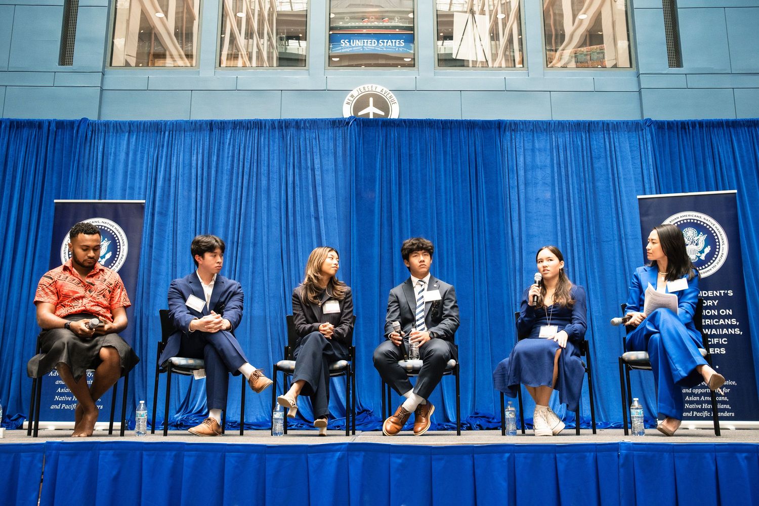 Six panelists seated on stage at a professional conference with blue curtains and banners in background.