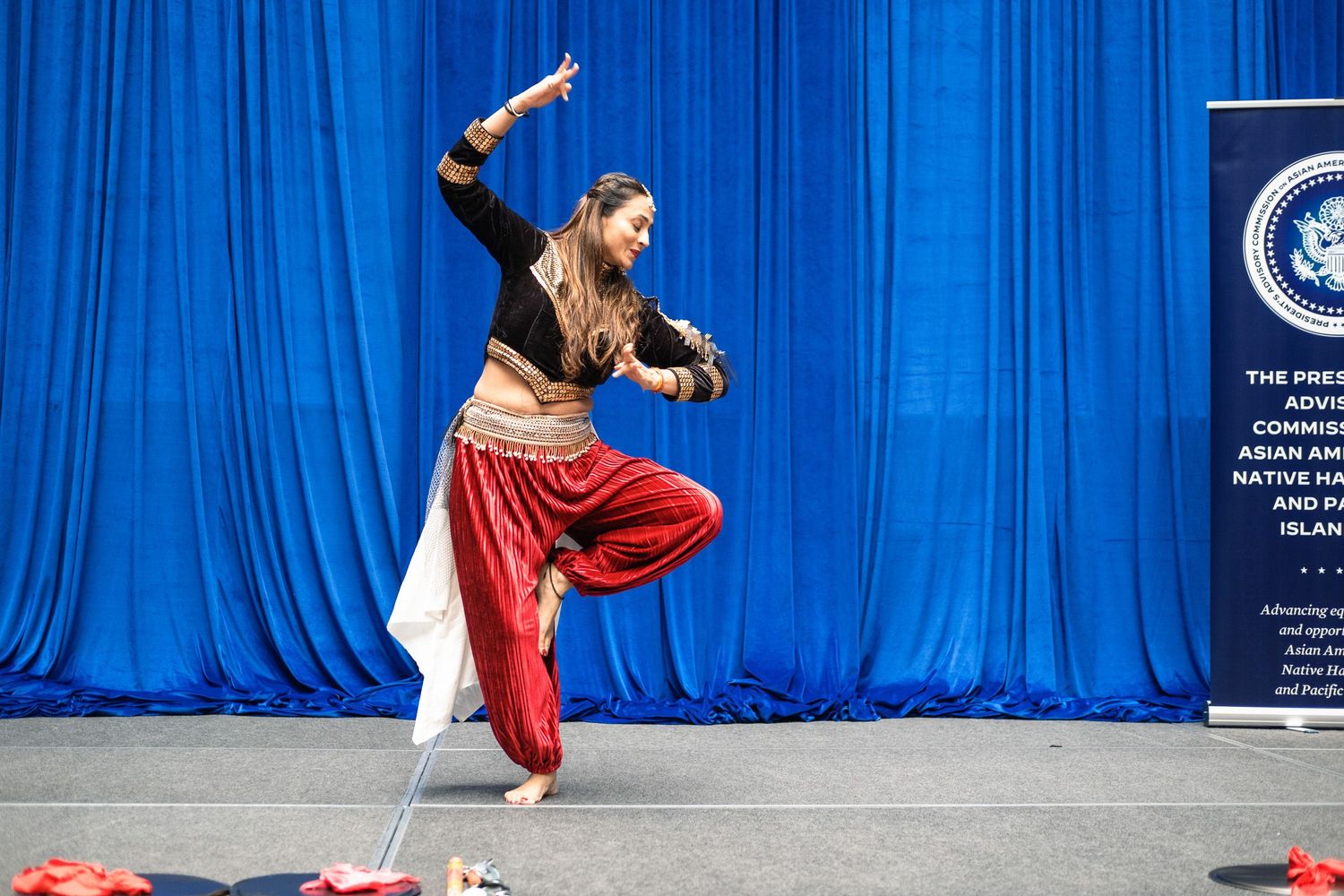 A woman performs a traditional Indian classical dance on stage with blue curtains and an official banner in the background.