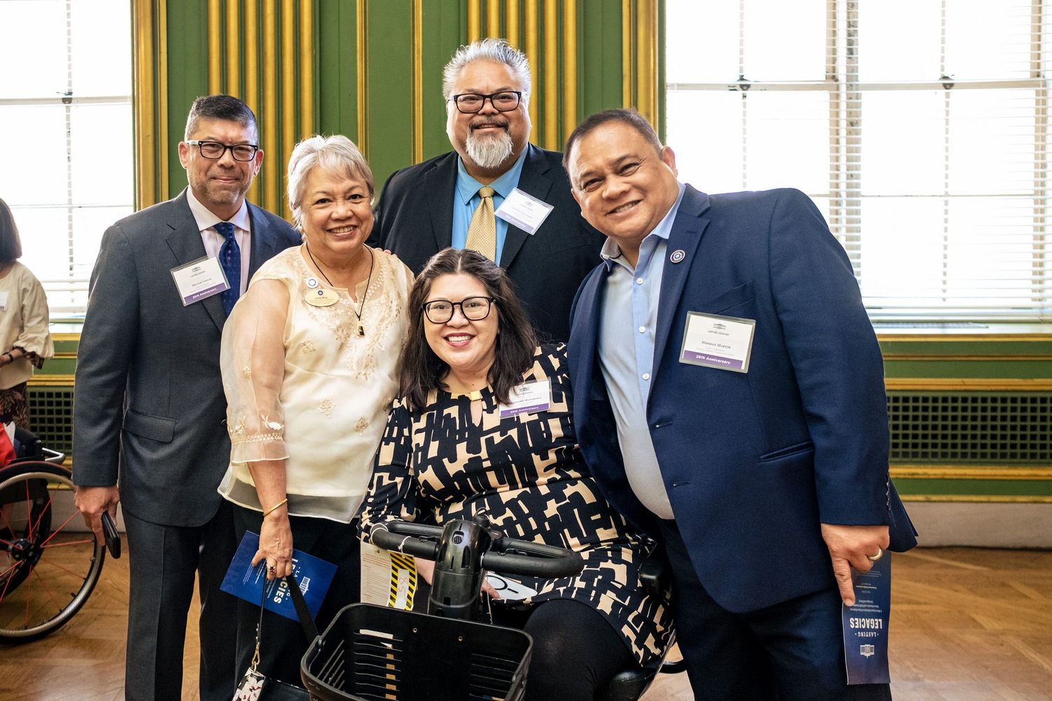 Five diverse professionals smile together at a formal indoor event, one seated in a wheelchair.