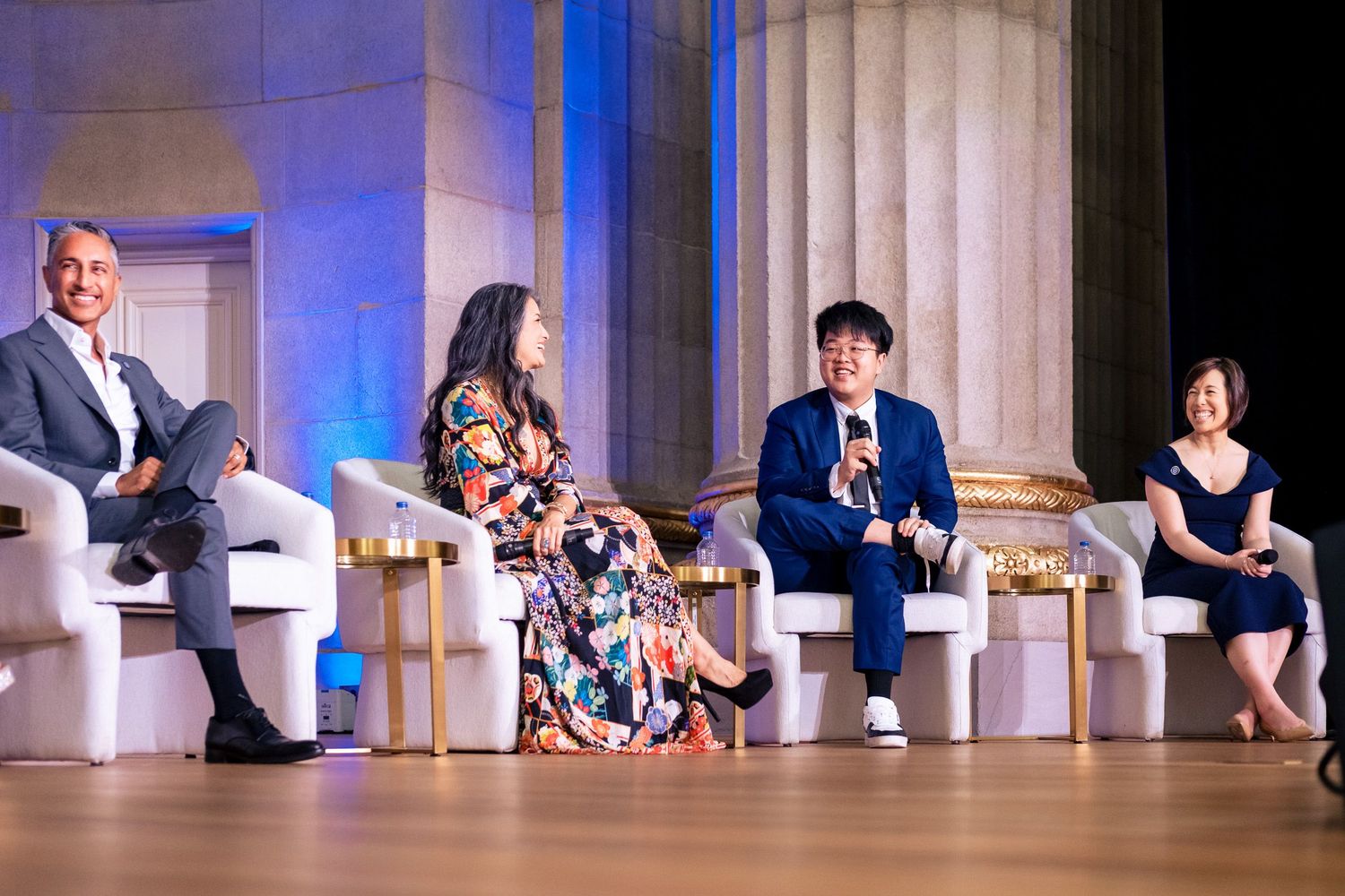 Four panelists seated on stage in white chairs at a formal event with classical columns in the background.