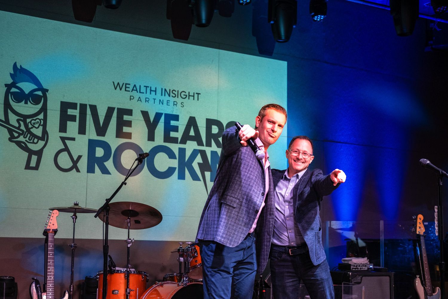 Two men celebrate on stage at Wealth Insight Partners Five Years & Rocky anniversary event with band equipment visible.