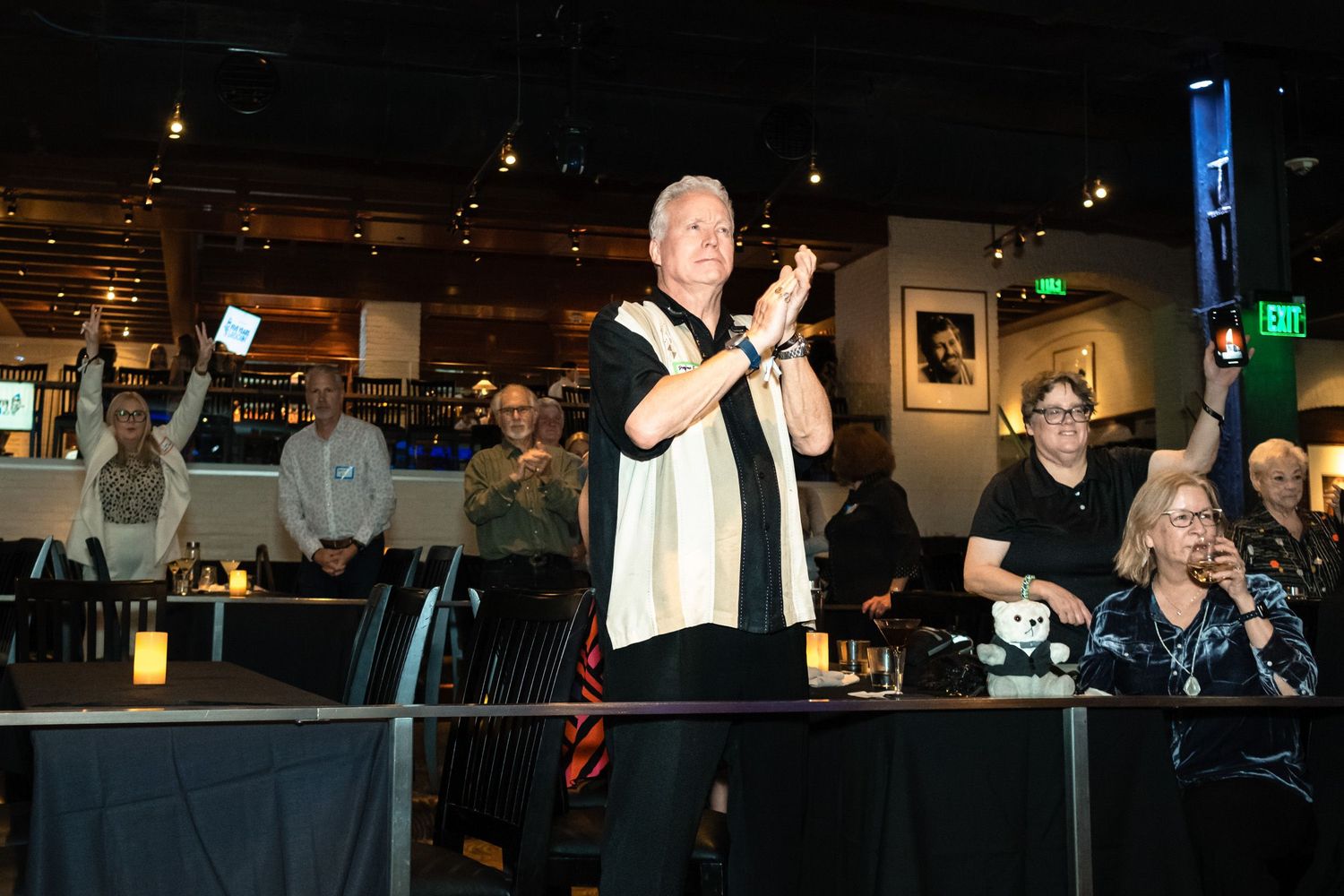 Man clapping at indoor event with audience raising hands in dimly lit venue.