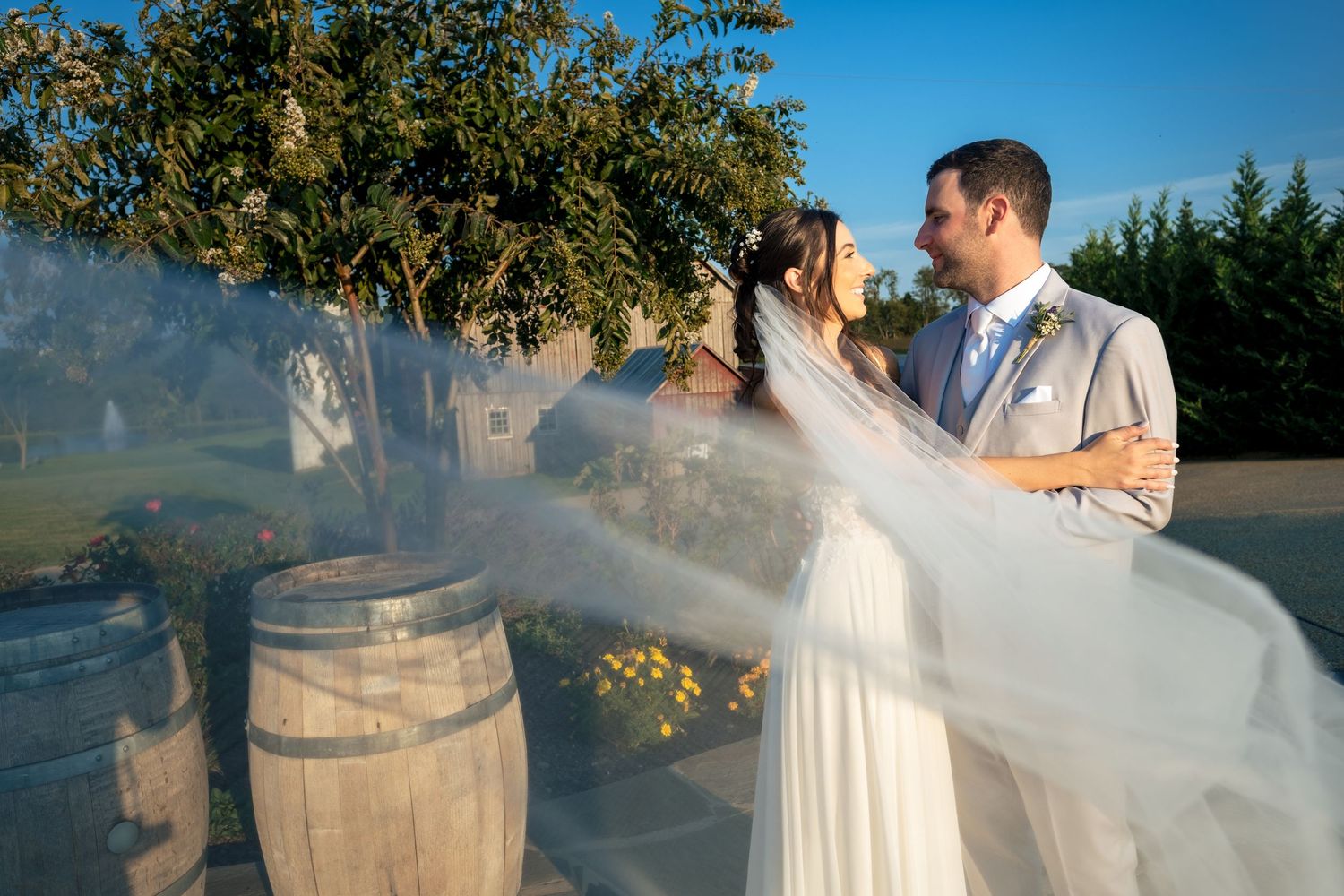 Bride and groom share a romantic dance outdoors near rustic wine barrels with a flowing veil in the breeze.