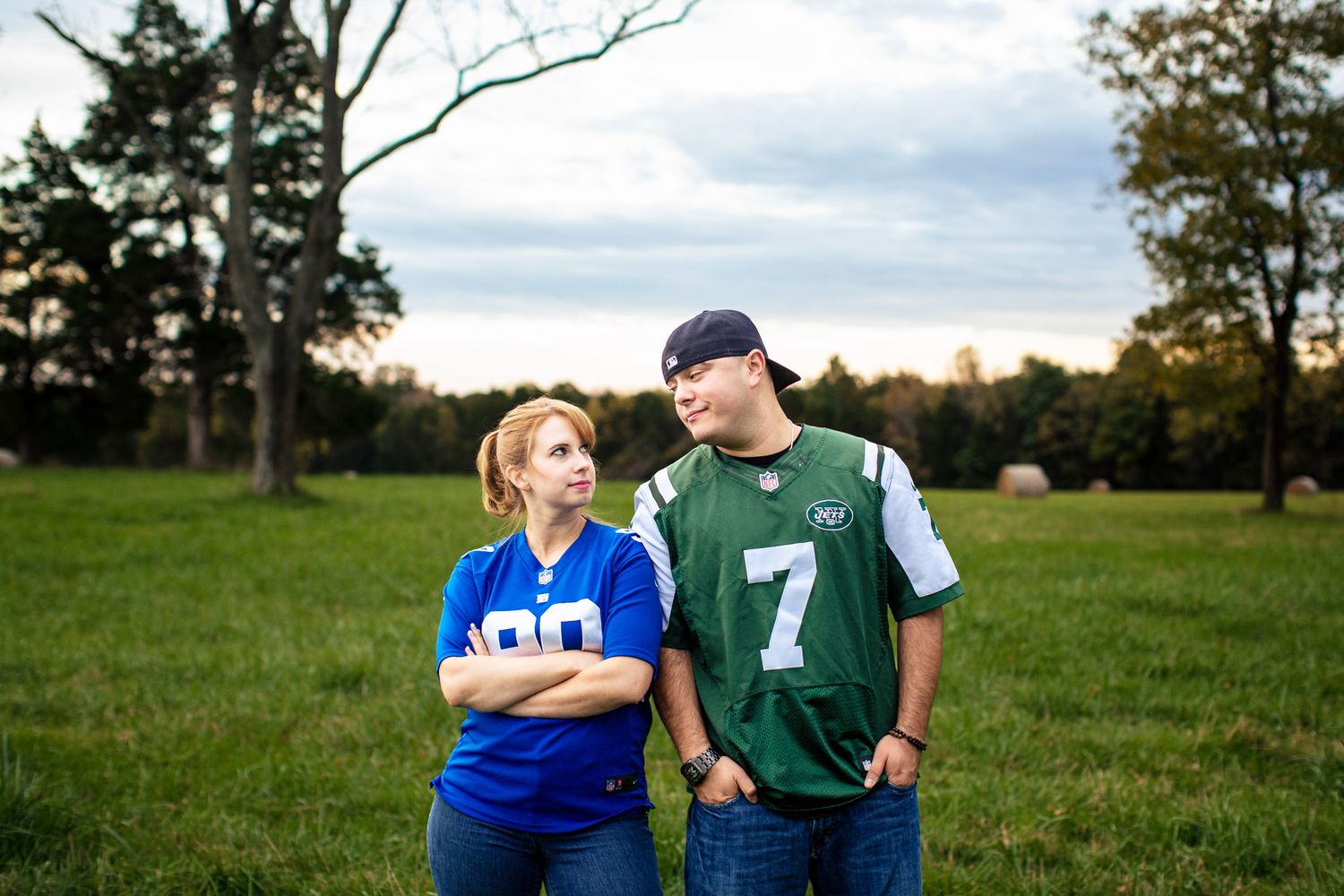 Couple in rival NFL team jerseys standing apart playfully in an open grassy field at dusk.