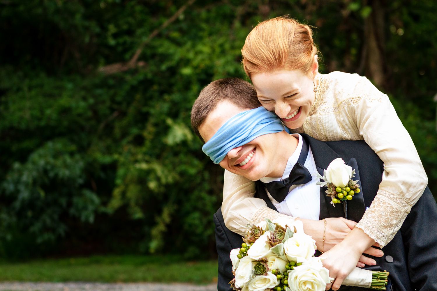 Playful bride blindfolds groom with blue ribbon while hugging him from behind in a lush green garden.
