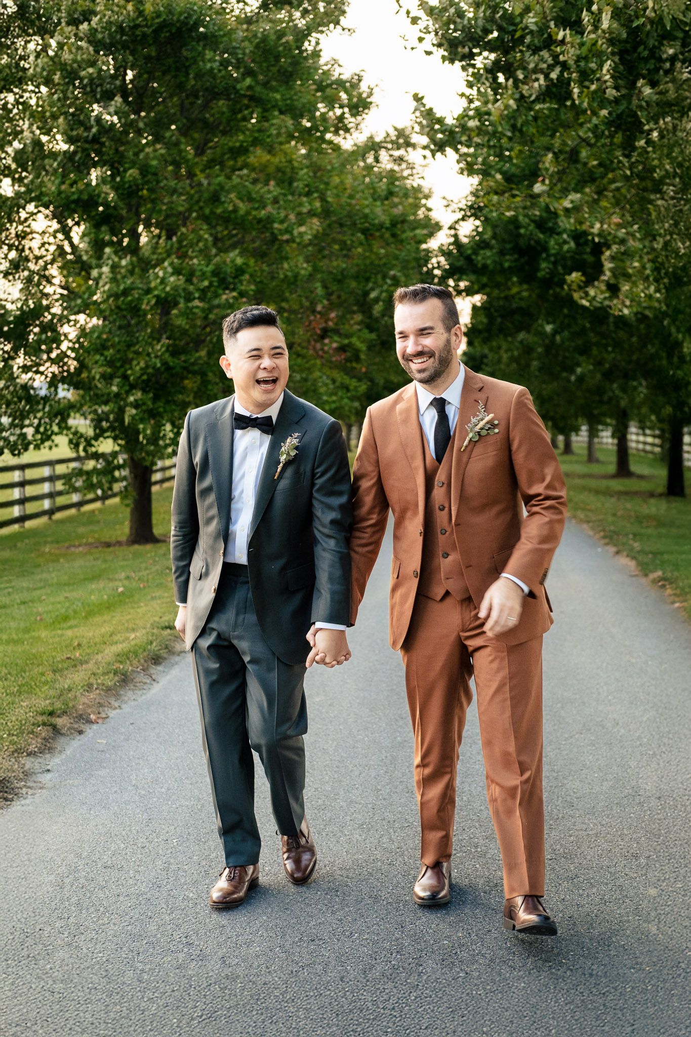 Two grooms in suits holding hands and laughing while walking down a tree-lined path.