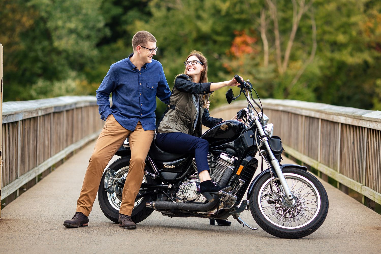 Couple smiling at each other beside a black motorcycle on a wooden bridge surrounded by autumn trees.