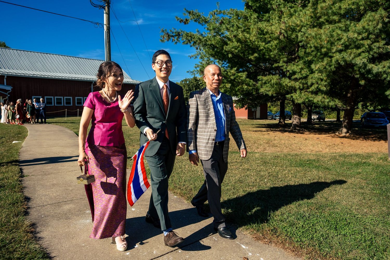 Two guests escort a man carrying a Thai flag along a sunny path at an outdoor wedding celebration.