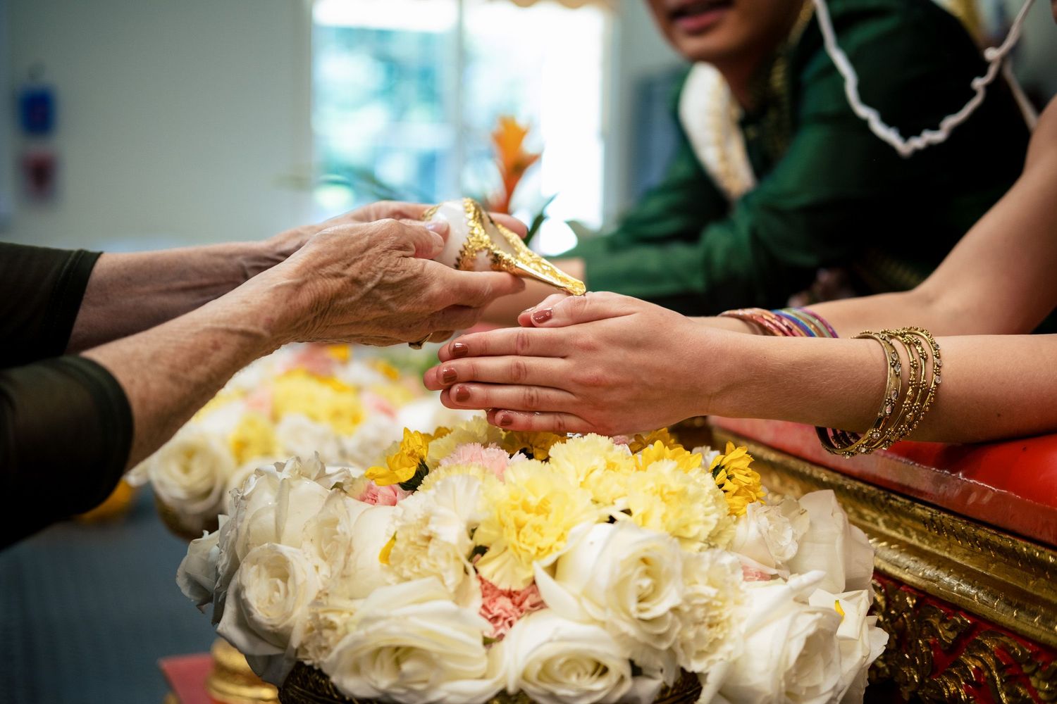 Elder hands blessing a couple during traditional Thai wedding water pouring ceremony with white flowers.