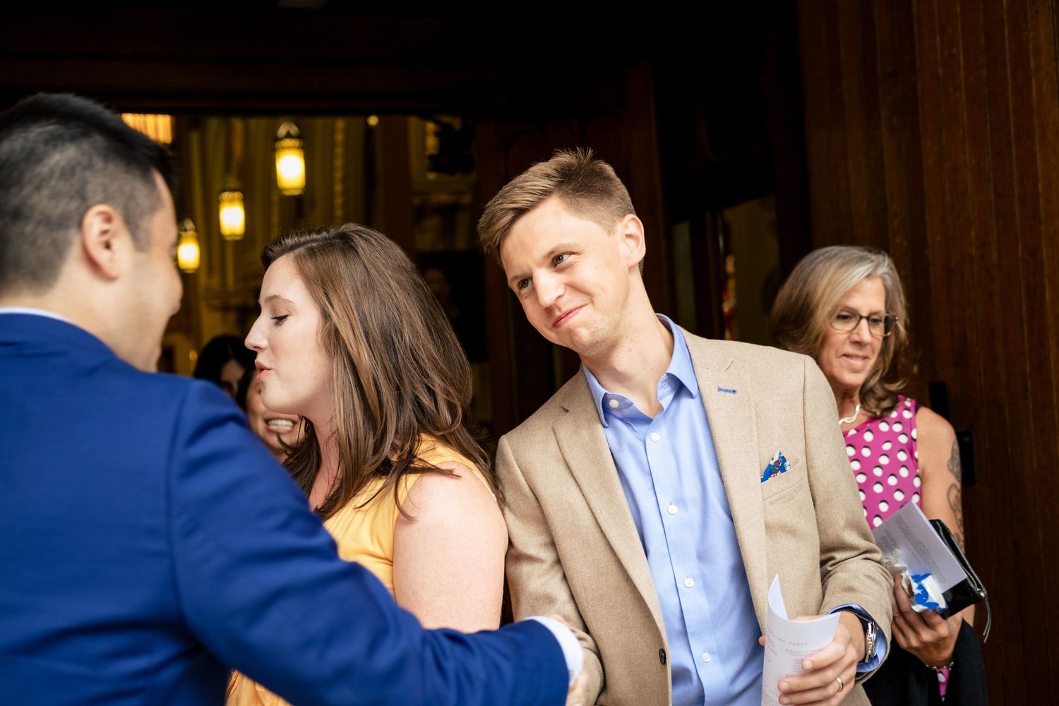 Wedding guests in tan blazer and yellow dress chatting with groom in blue suit at warmly lit venue entrance.