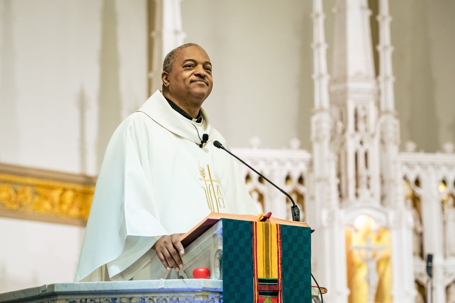 Priest in white vestments smiles warmly while delivering a wedding homily at the church altar.