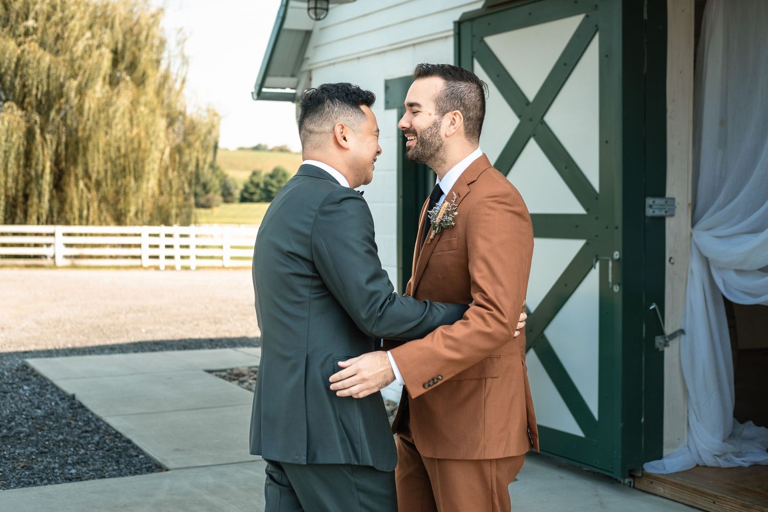 Two grooms sharing an emotional first look embrace outside rustic barn wedding venue.