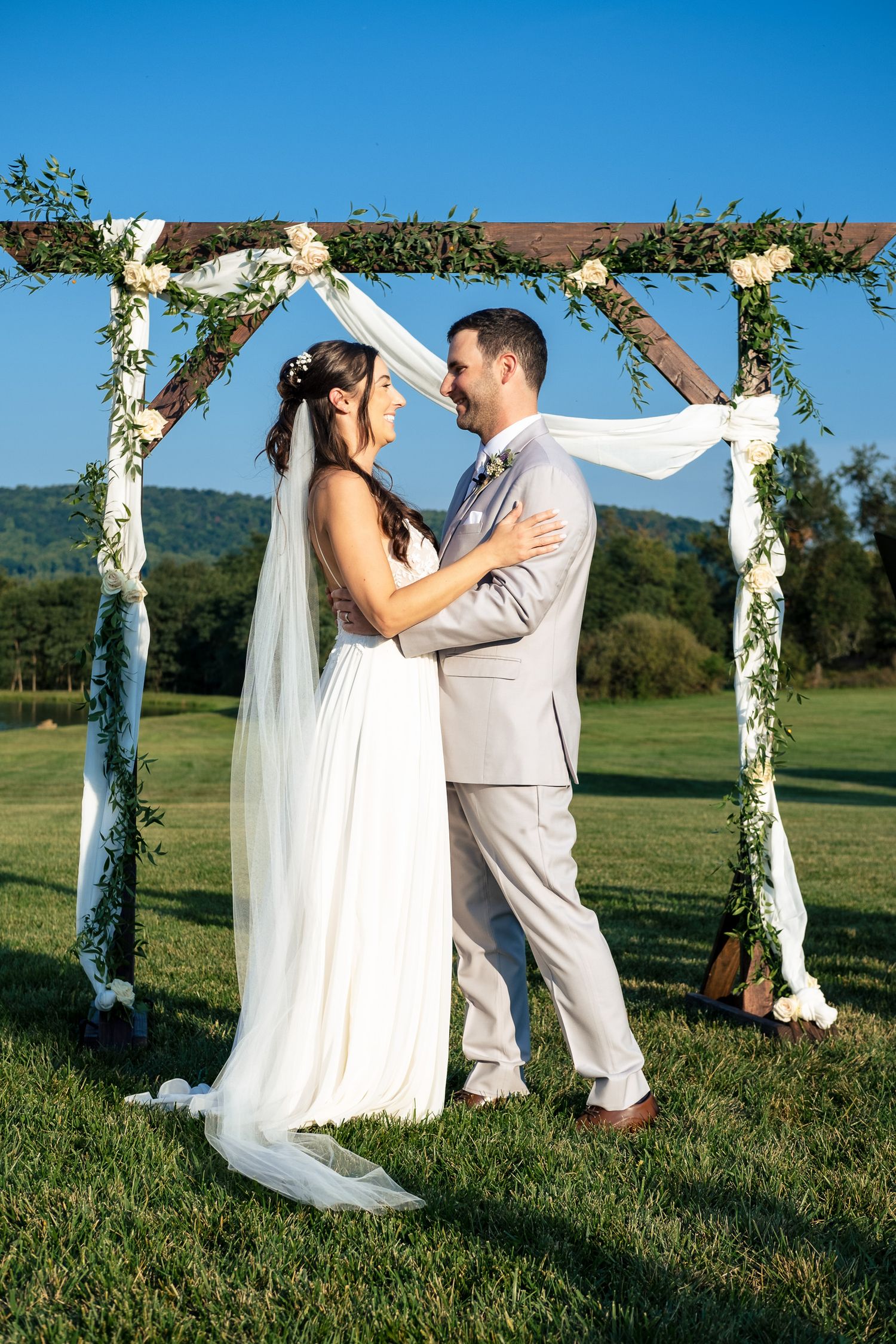 Bride and groom share a tender moment under a floral wooden arch on a sunny outdoor wedding day.