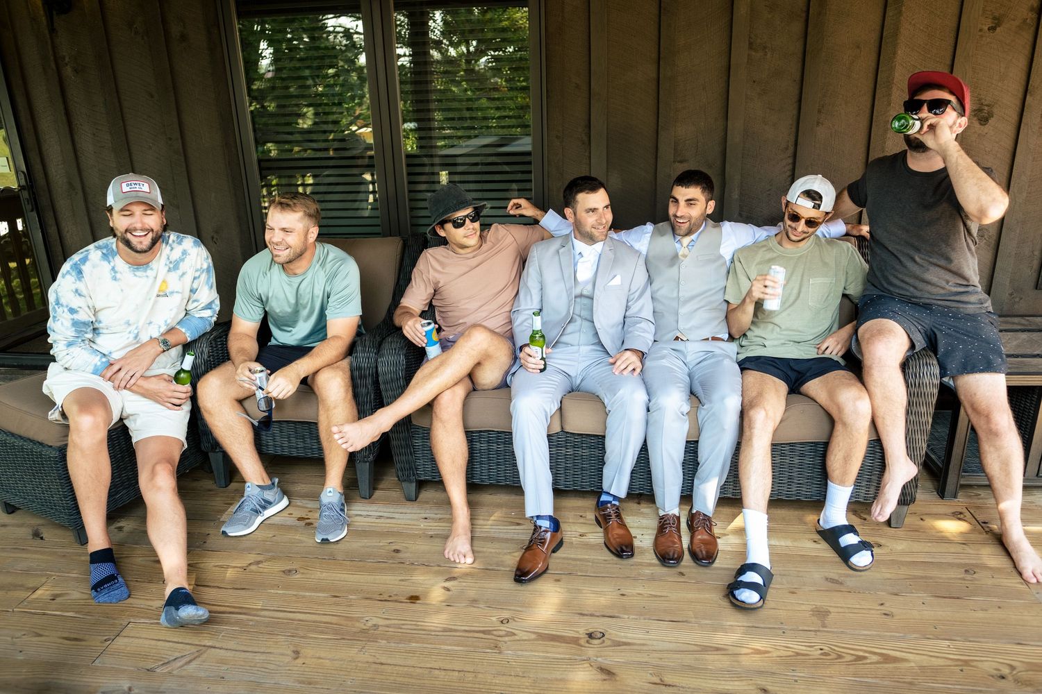 Groomsmen and friends casually dressed sitting together on a wooden deck bench outdoors.