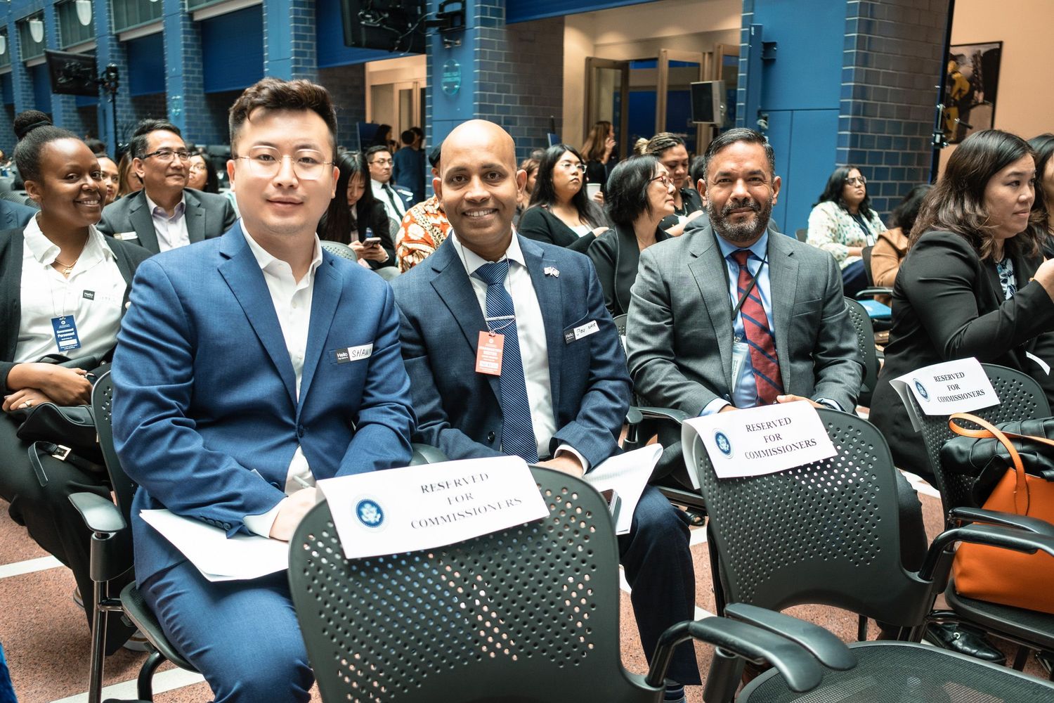 Three professionally dressed men smile while seated at a formal event, with name placards and attendees visible in the background.