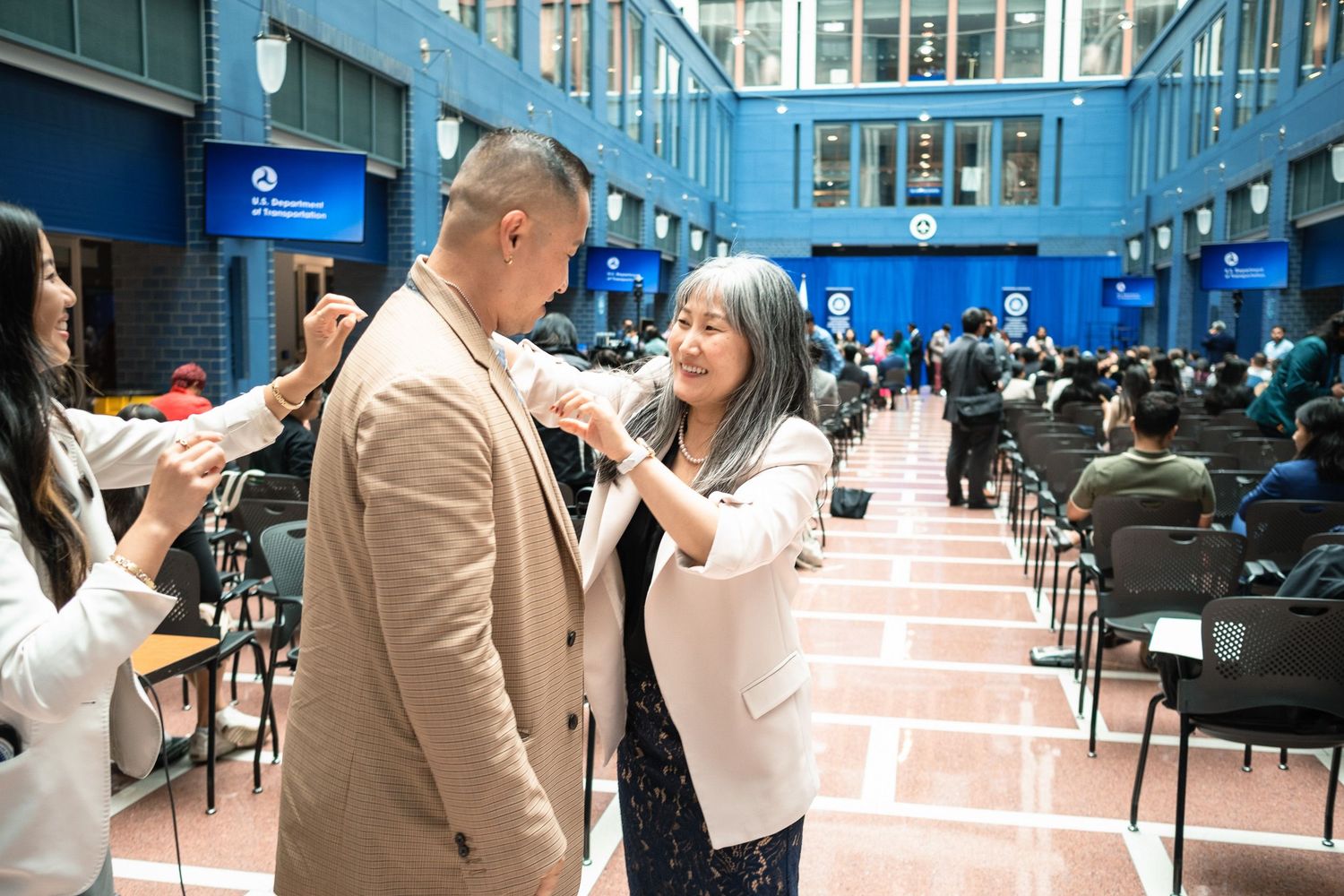 Woman in white blazer pins a corsage on a man in a tan suit at a formal ceremony.