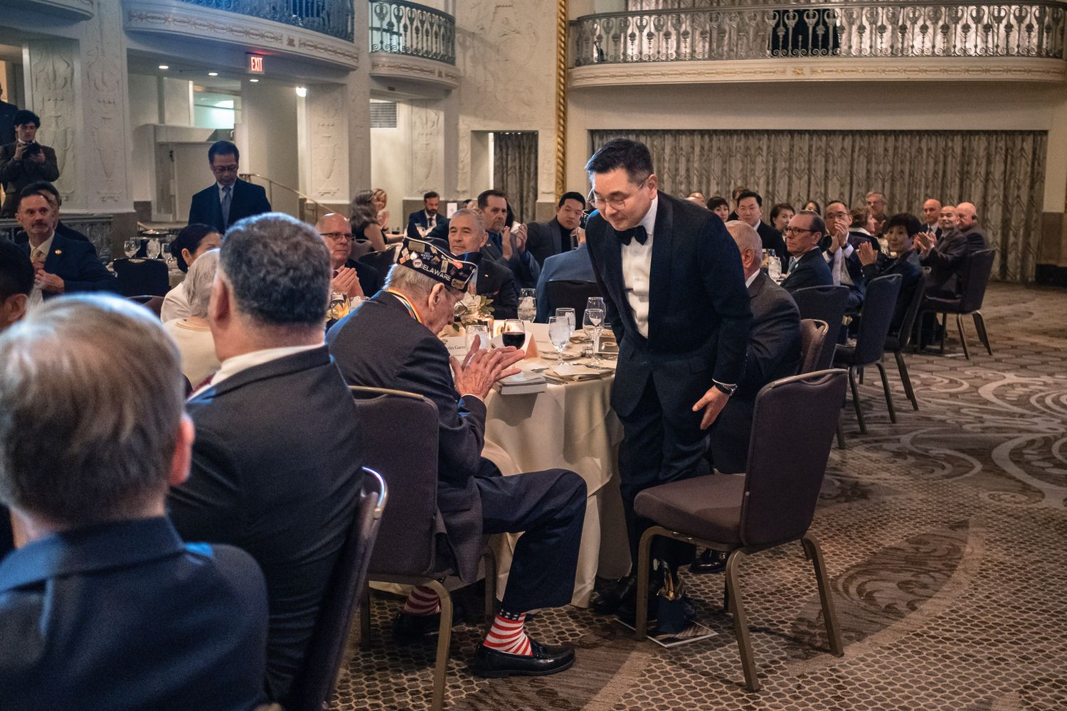 Man in tuxedo greeting seated guests at formal Korean War Veterans Memorial Foundation dinner.