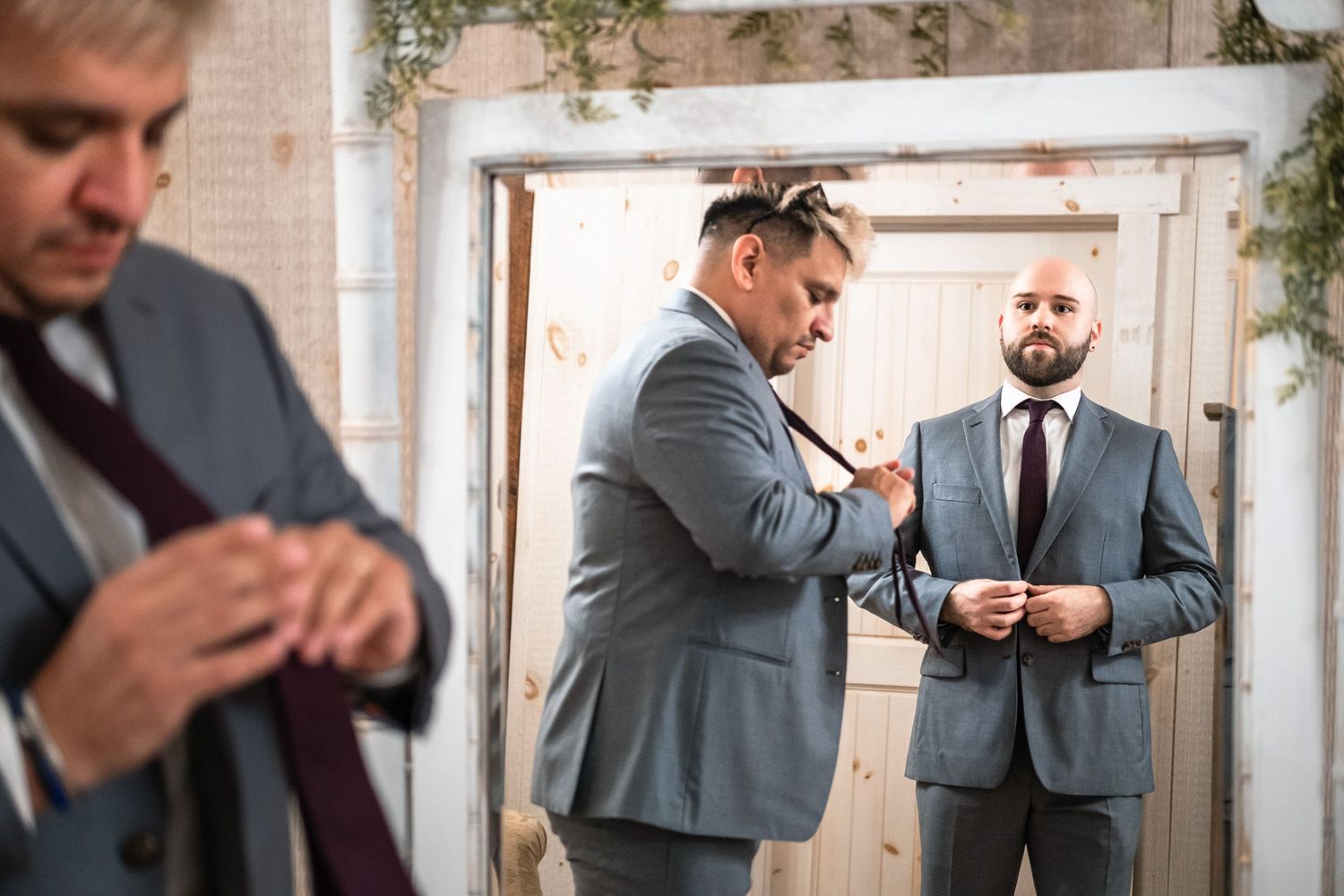 Groomsmen in gray suits adjusting ties and buttoning jackets in a rustic wedding venue.