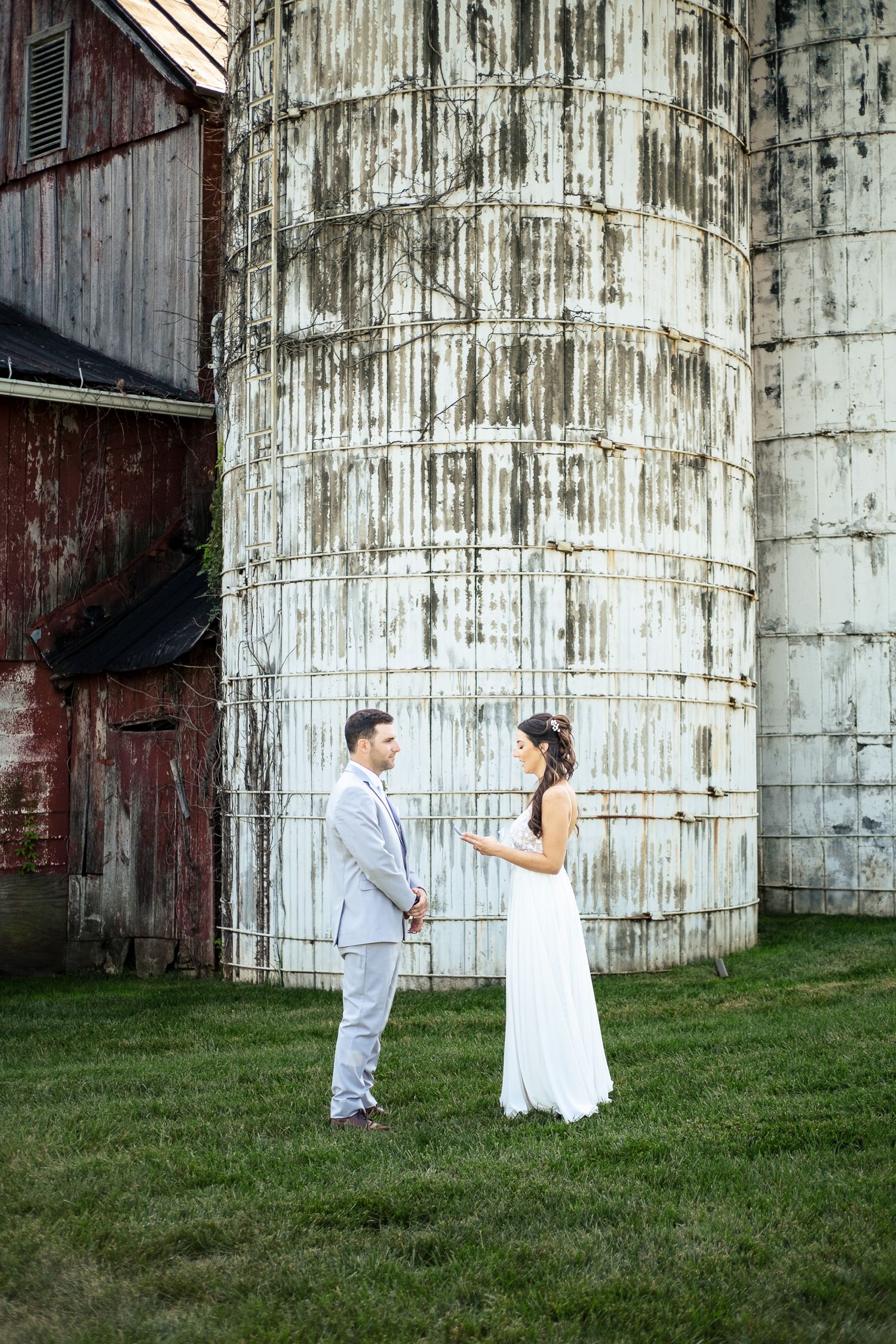 Bride and groom hold hands facing each other beside towering weathered white grain silo at farm.