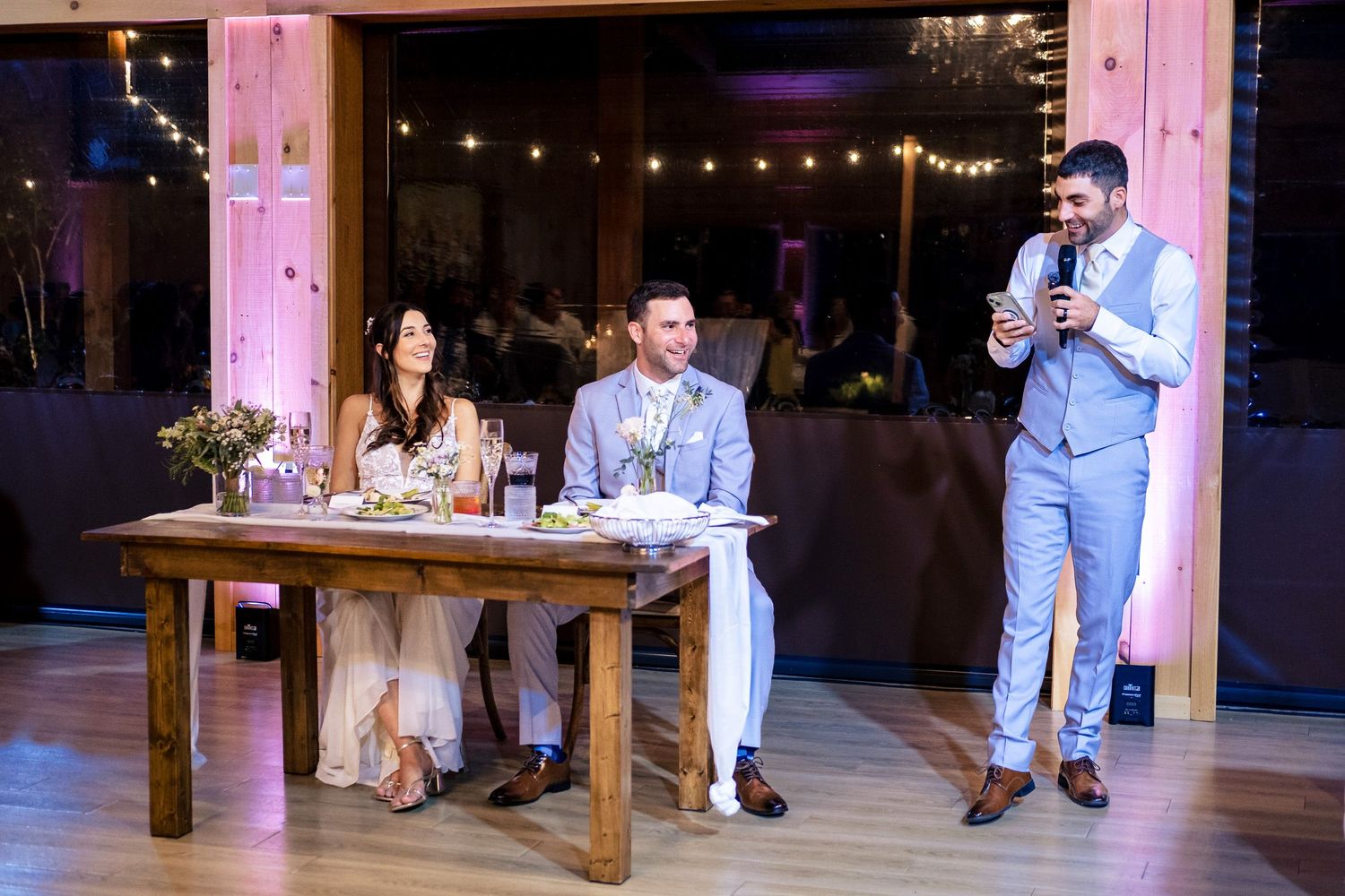 Best man delivering wedding toast while bride and groom laugh at head table.