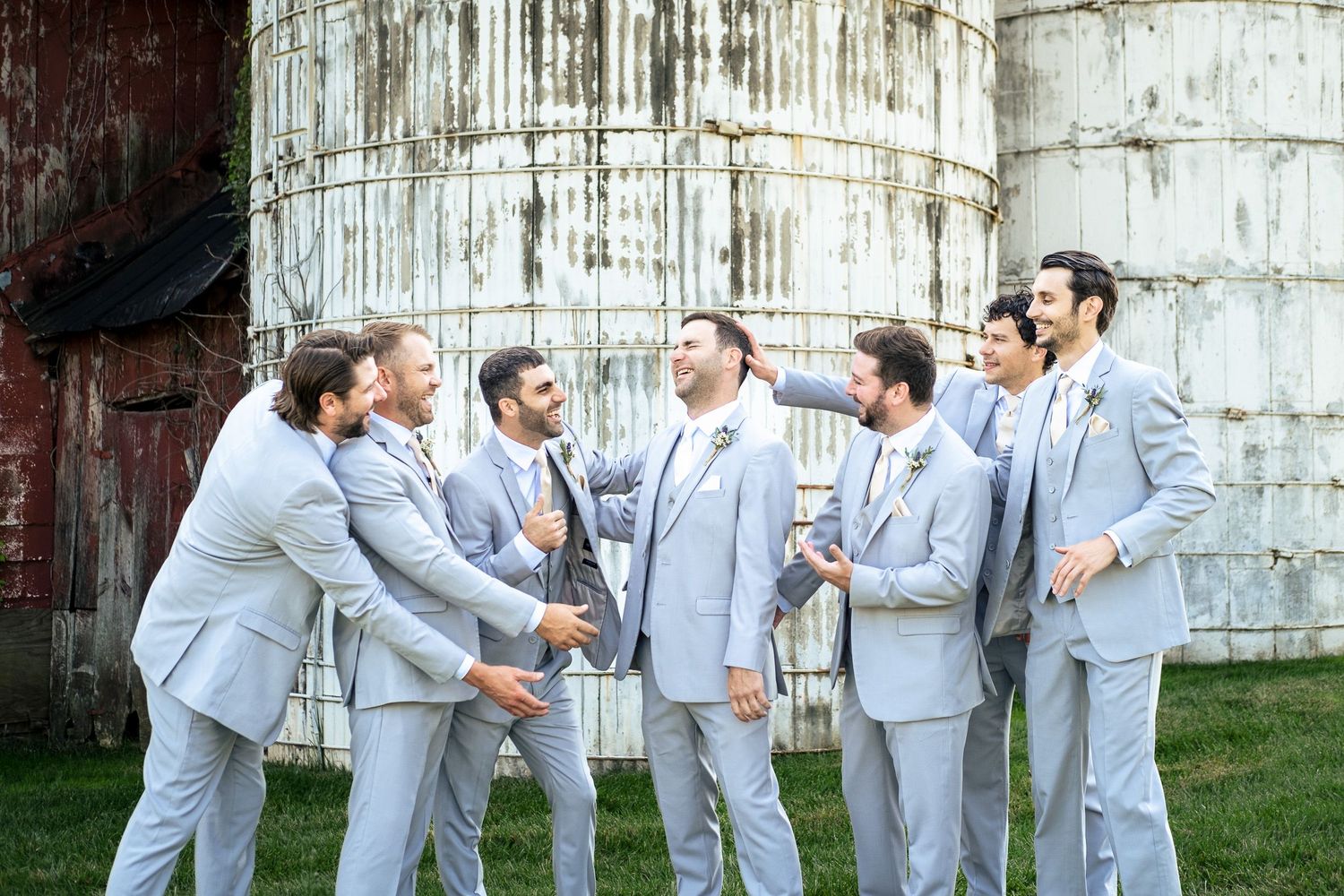 Groomsmen in light gray suits laugh together near weathered grain silos at a farm wedding.