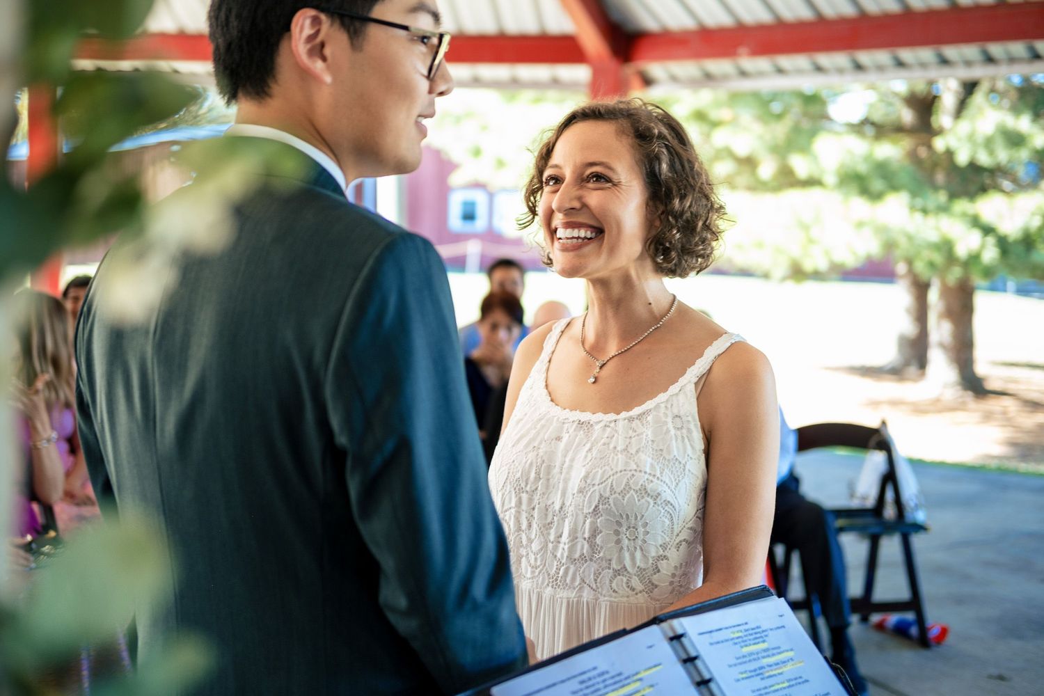 Radiant bride smiling at groom during outdoor wedding ceremony under a red pavilion roof.