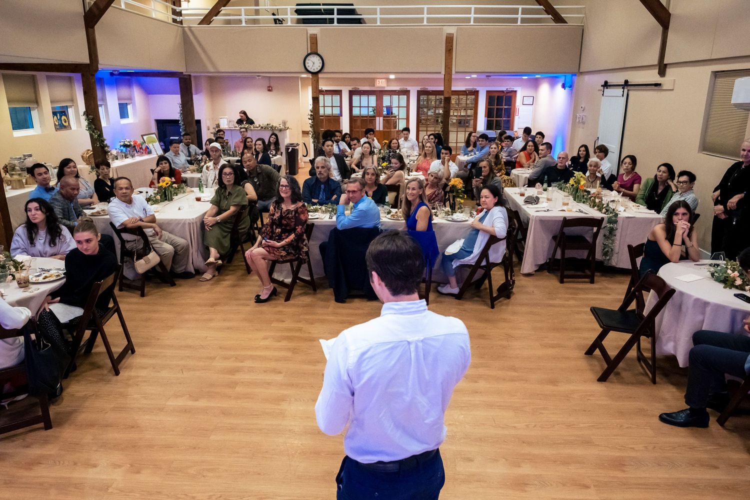 Man in white shirt addresses guests seated at round tables during a wedding reception in a rustic hall.