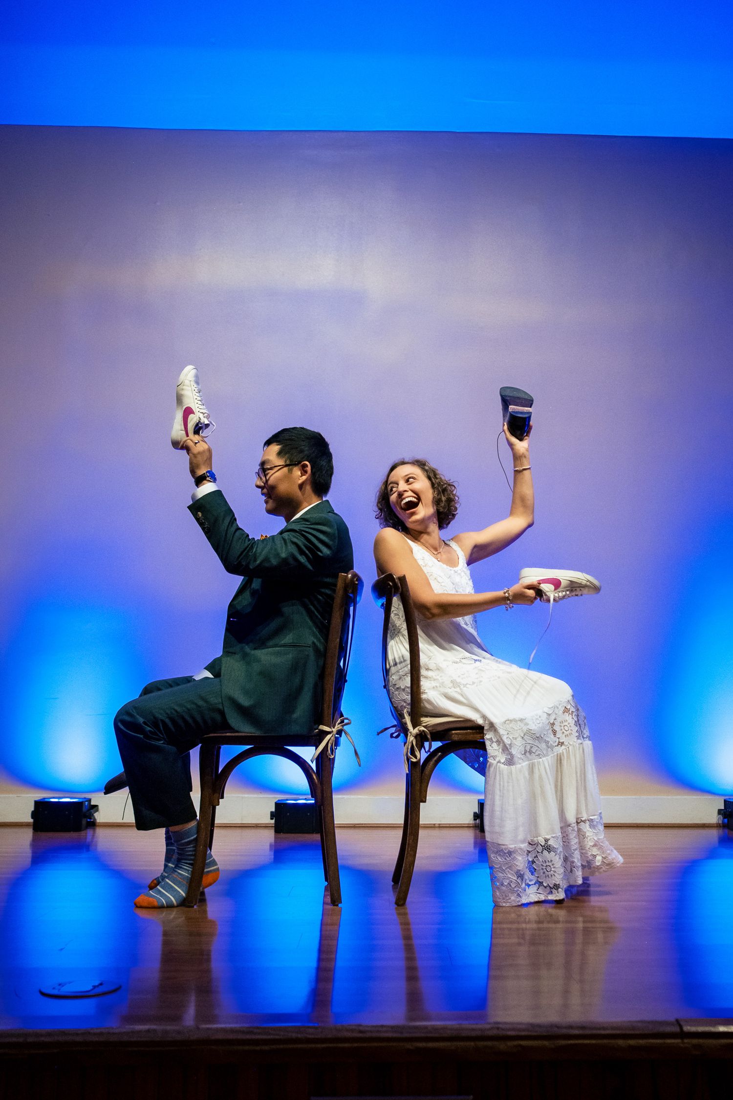 Bride and groom laugh while holding up shoes during fun wedding shoe game on stage.