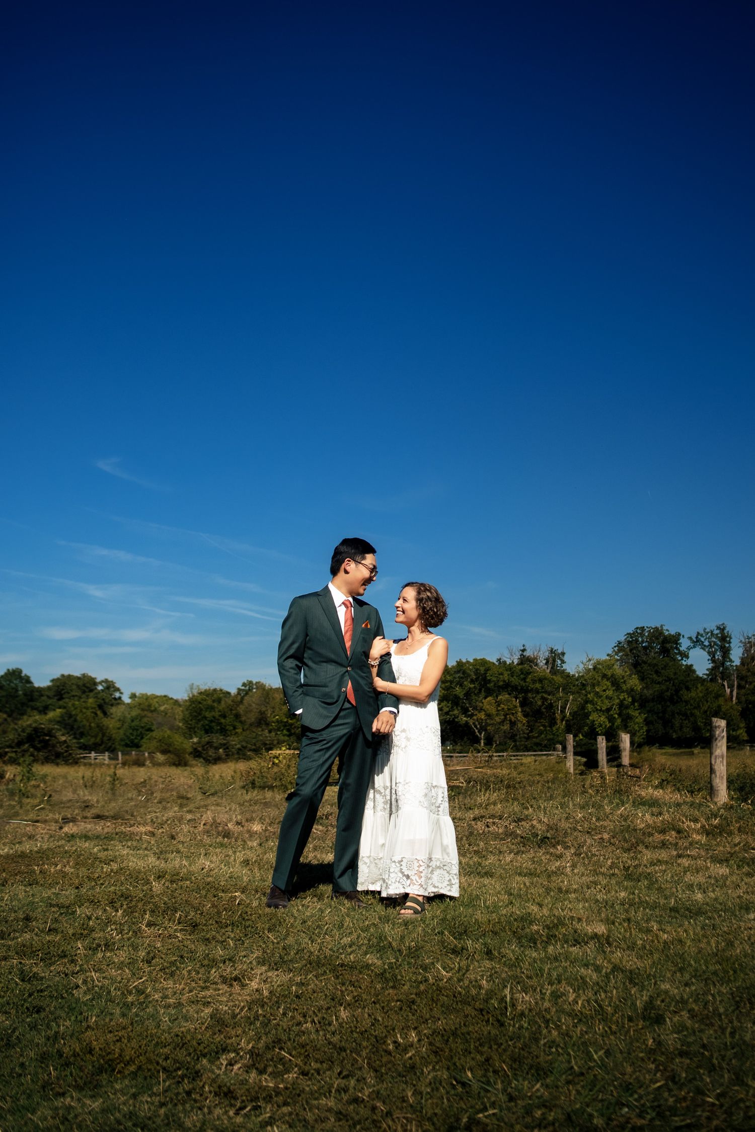A bride and groom share a joyful moment together in a green field under a deep blue sky.